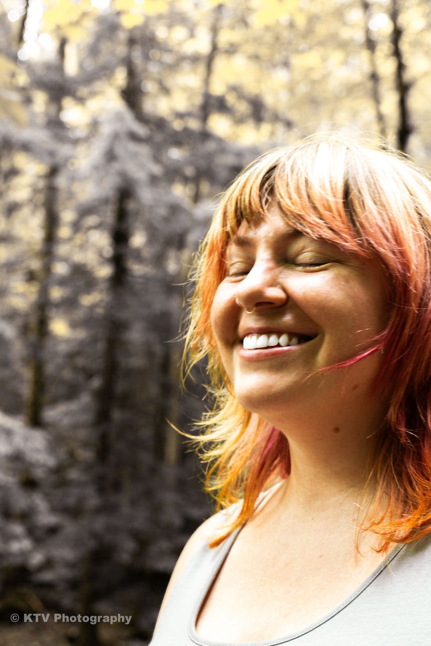 Smiling woman with red and pink hair enjoying nature in an autumn forest, eyes closed in the sunlight.