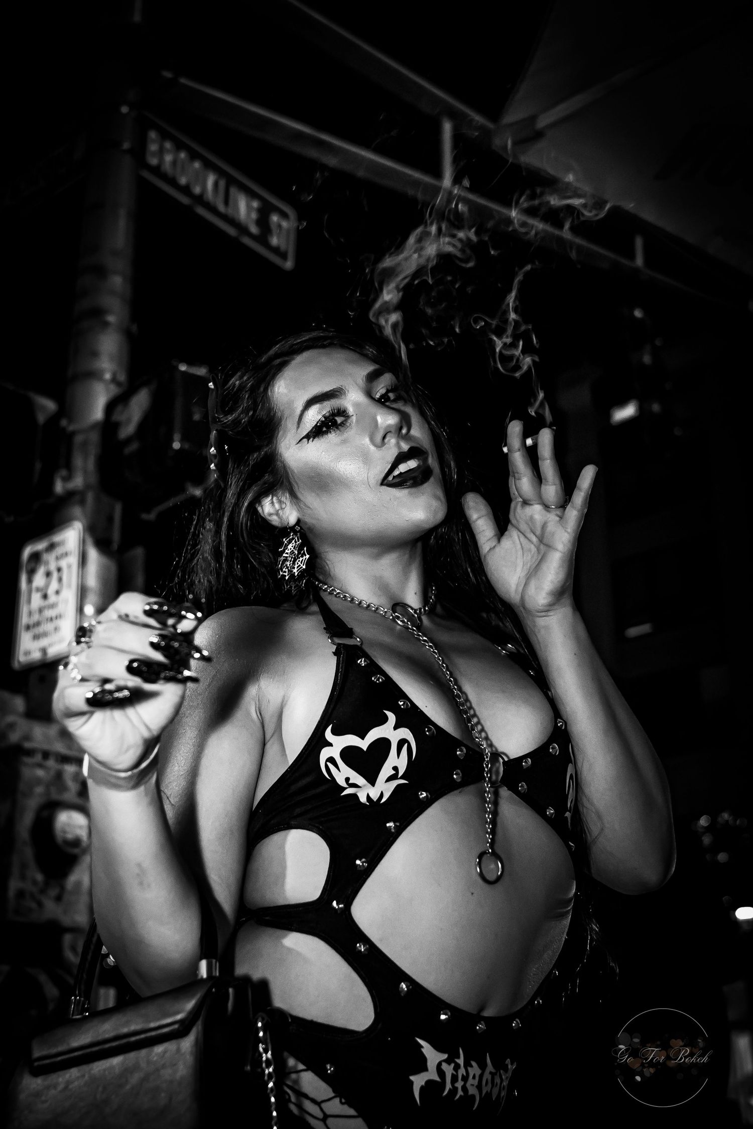 Black and white photo of a woman in gothic outfit smoking dramatically on a city street at night.