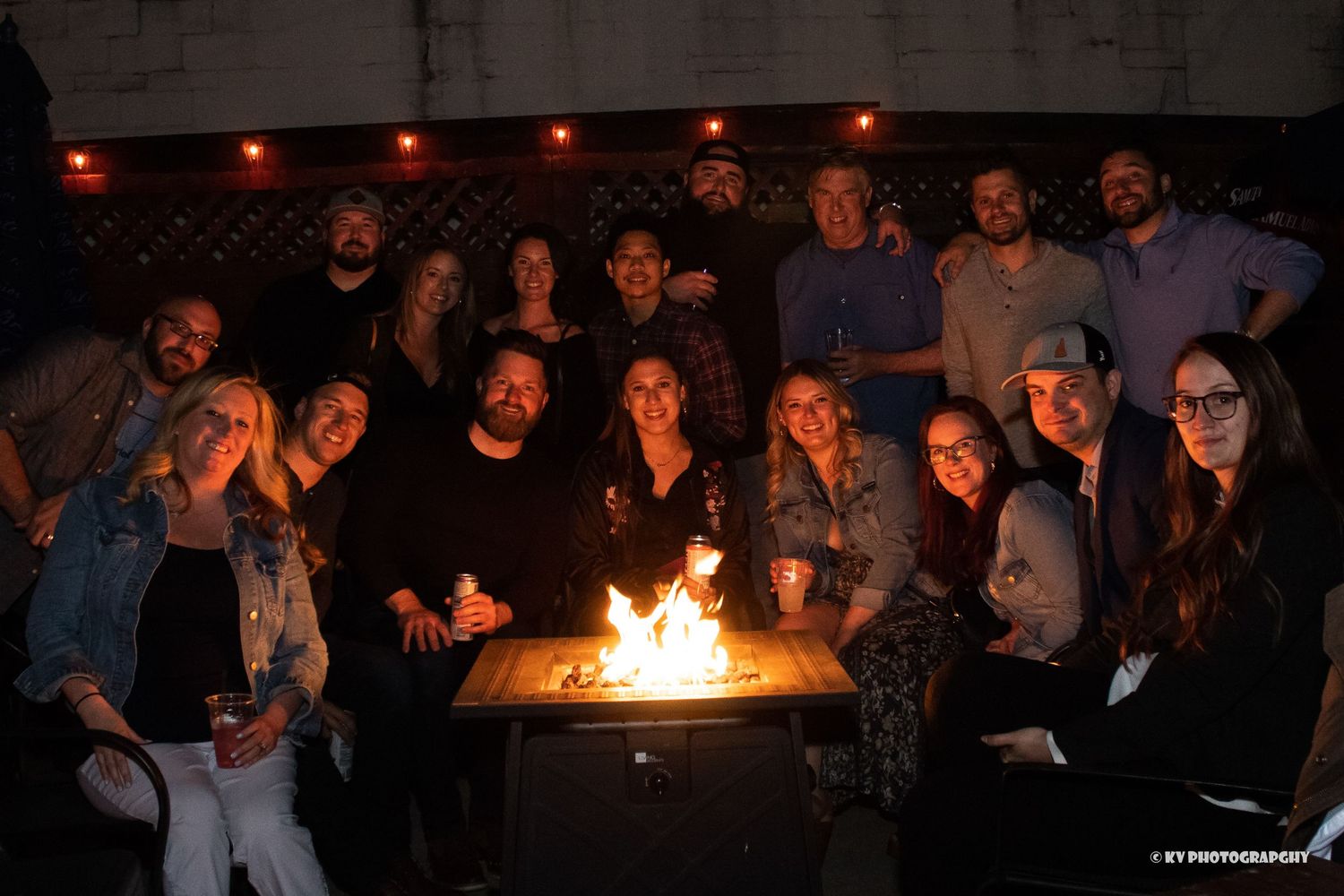 Group of smiling friends gathered around an outdoor fire pit at a nighttime social event.