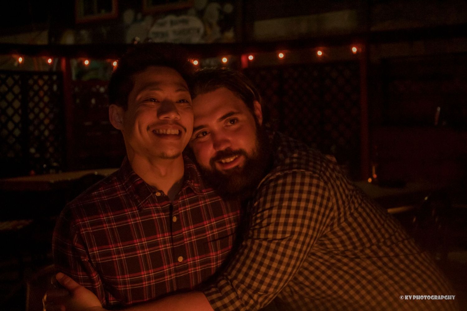 Two bearded men smiling and posing together in a warmly lit bar setting.