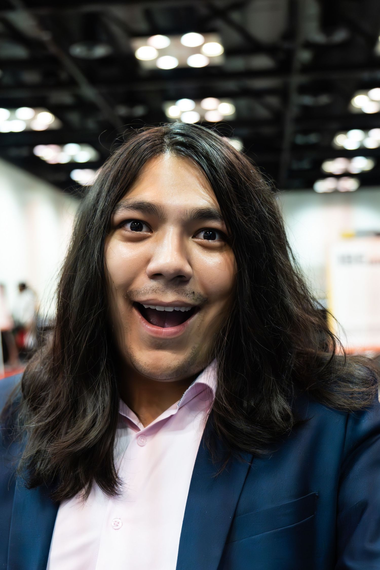 Young man with long dark hair and braces smiling at a convention center.