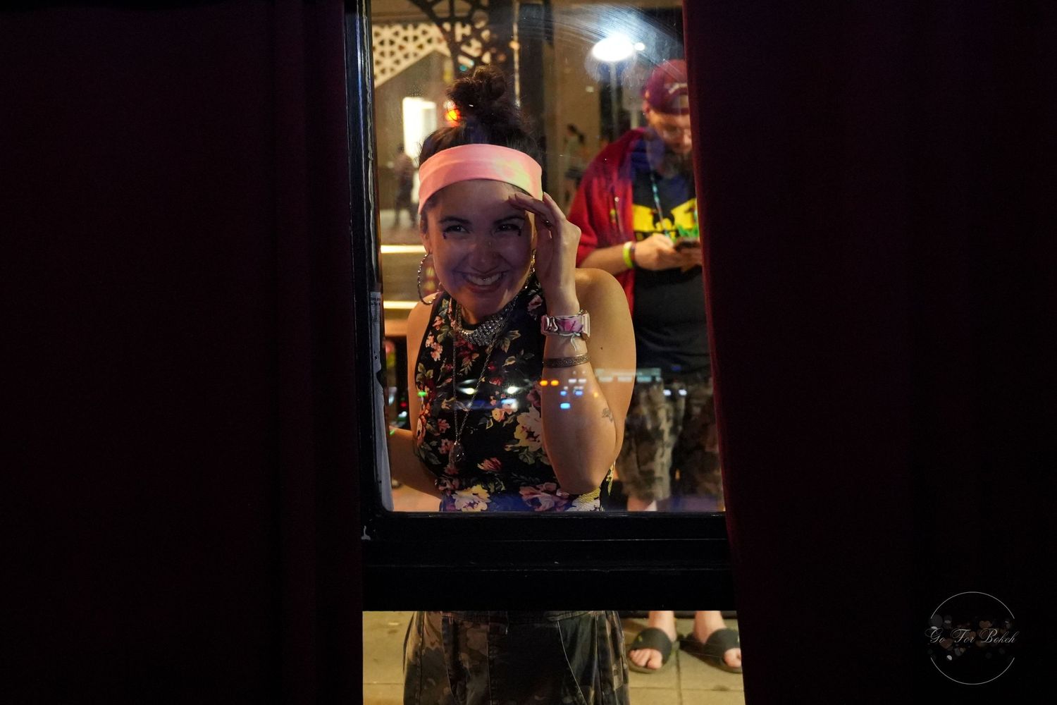 Smiling woman with pink headband and colorful beads looking through a window at a night event.