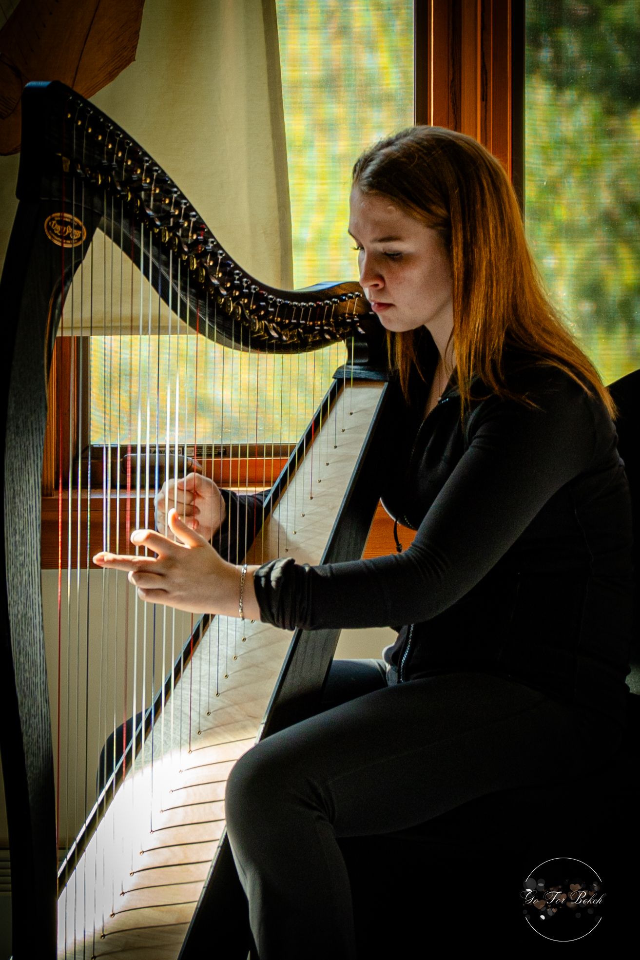 Young woman with red hair playing a black concert harp by a sunlit window indoors.