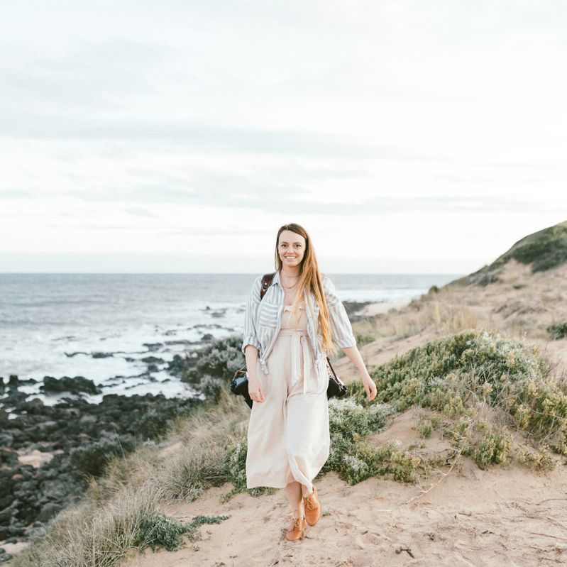 Woman in flowing dress walks barefoot on coastal trail overlooking rocky shoreline.