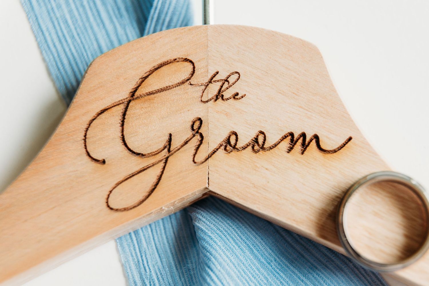 Wooden hanger engraved with 'The Groom' text and wedding band against blue fabric.