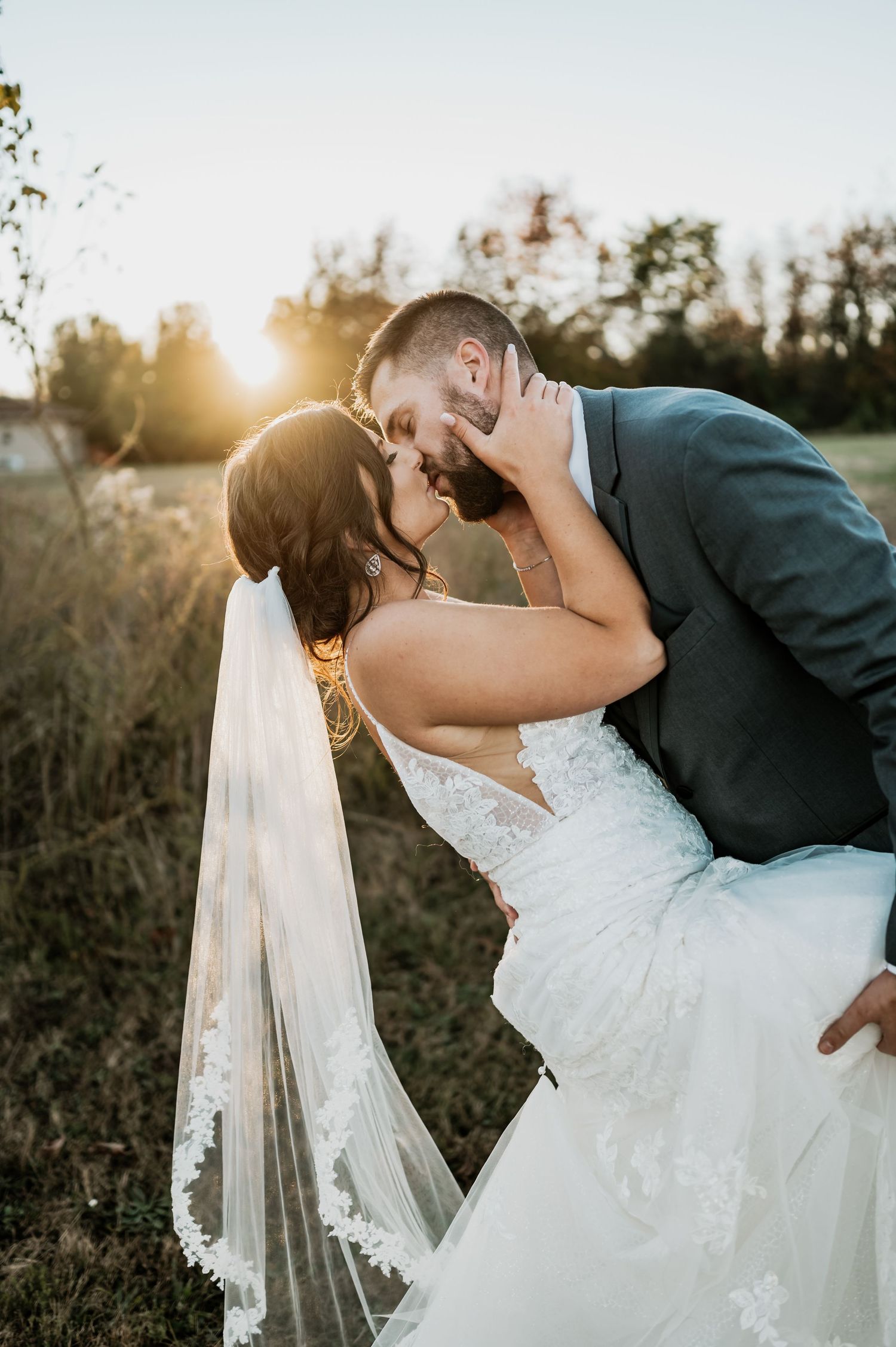 A romantic sunset wedding photograph of a couple sharing an intimate kiss in a rustic outdoor field setting.