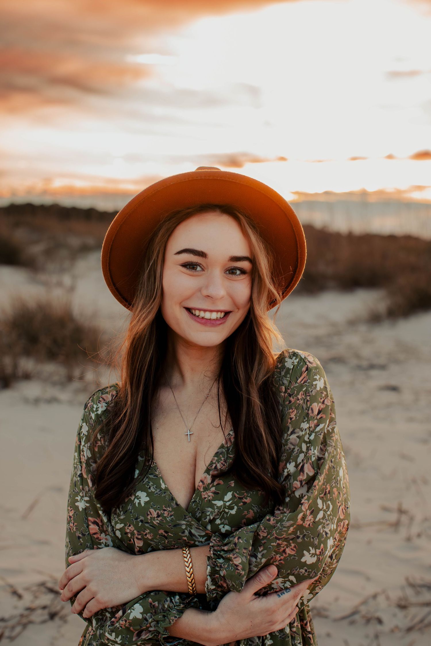 Woman wearing brown hat and floral dress poses against sunset in desert landscape.
