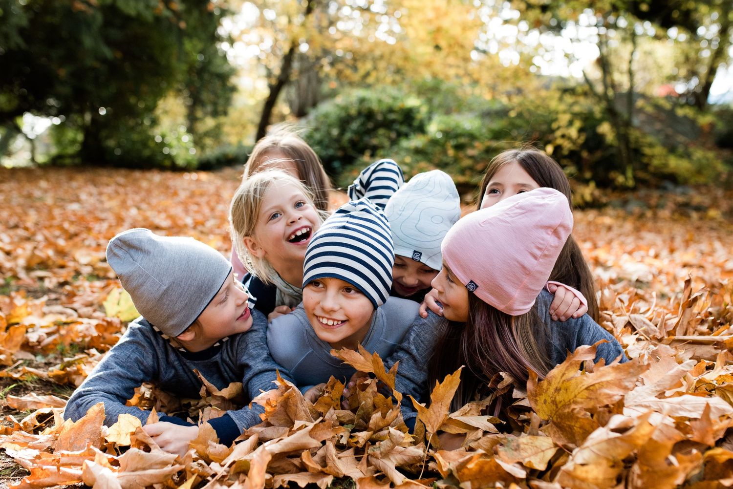 A bunch of children lying in a pile of leaves in the fall with hats on.