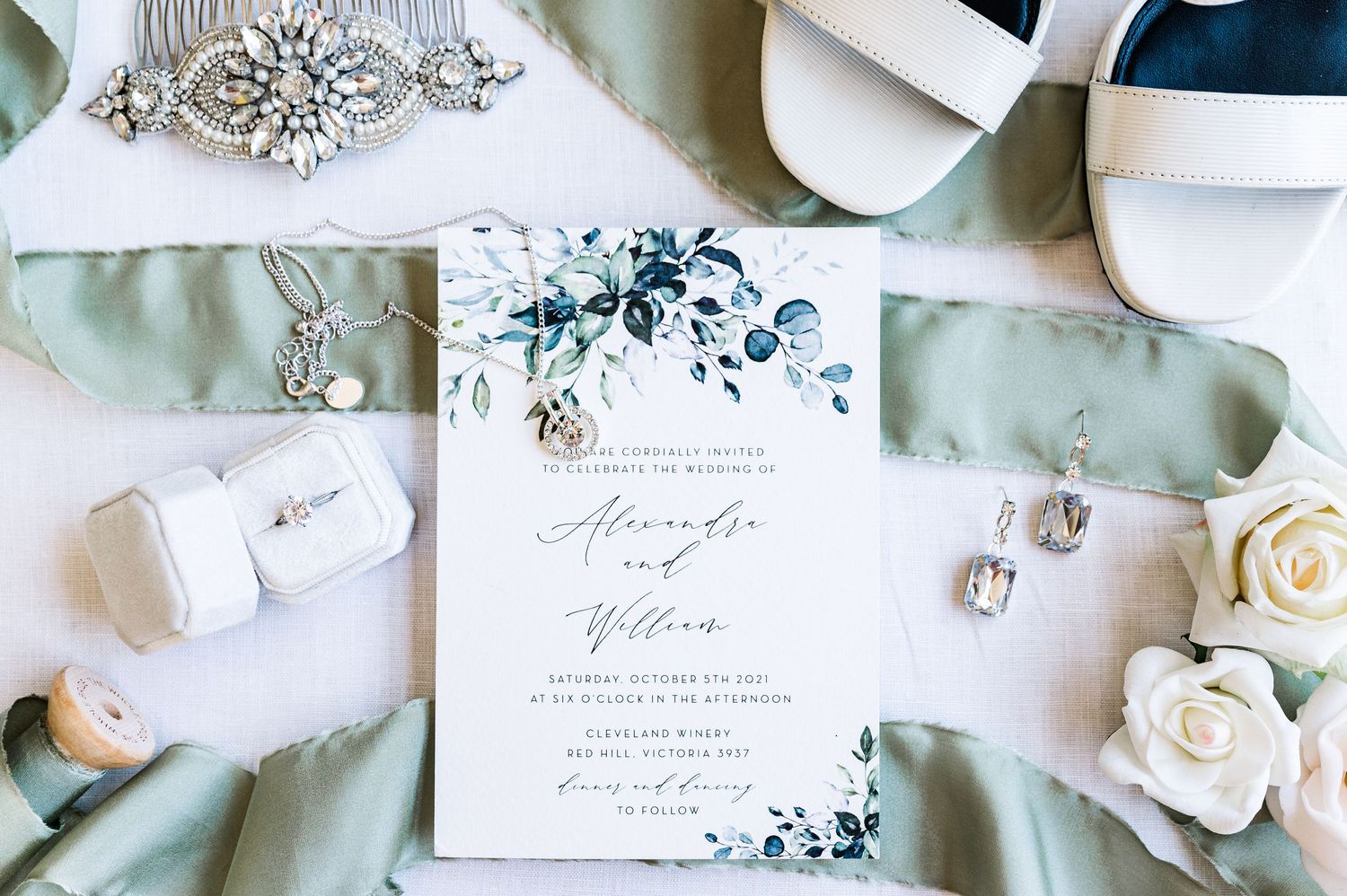 Wedding invitation with sage green styling, floral details and elegant accessories on light fabric.