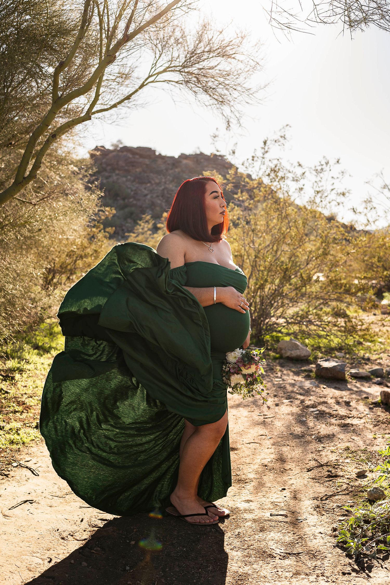 A pregnant women wearing an elegant green dress that is billowing behind her in the desert at South Mountain in Phoenix