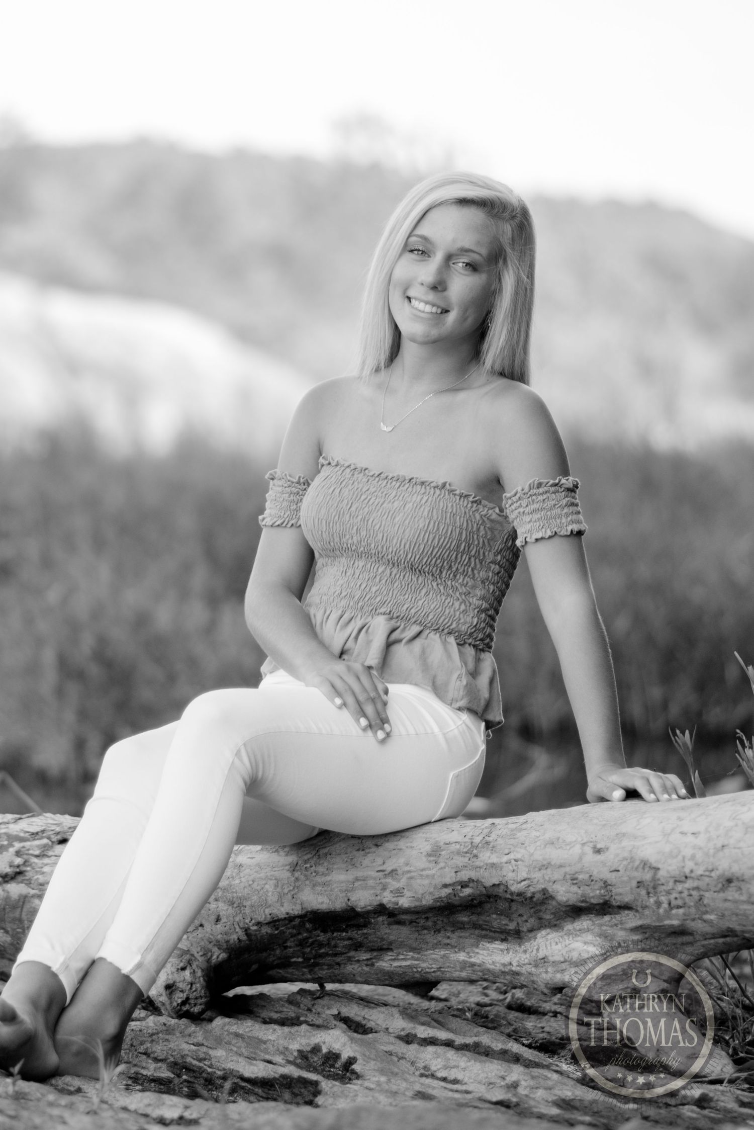 Woman in casual attire sits on a fallen log during a black and white outdoor photo session.