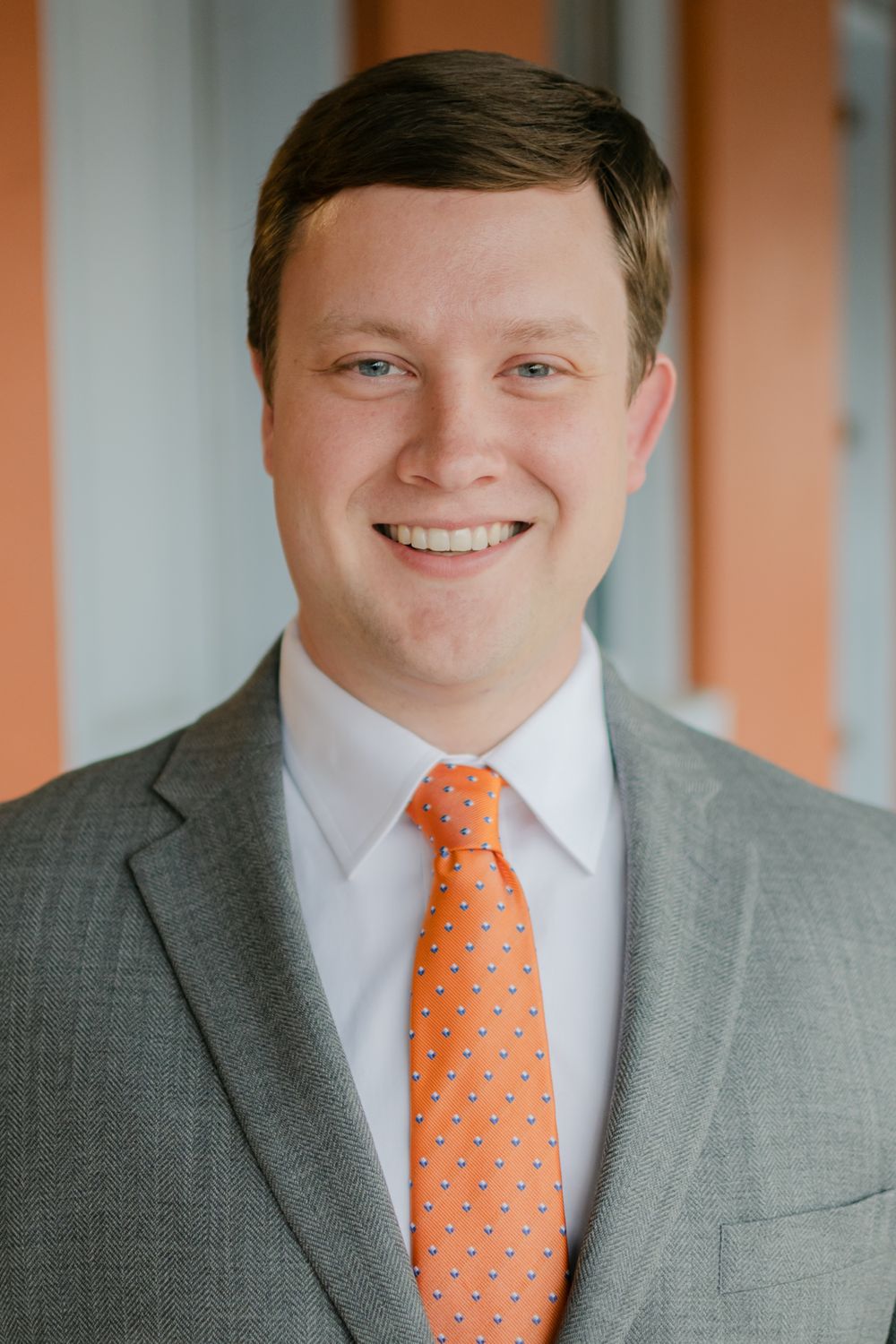 Professional headshot of a young man in a gray suit and orange tie smiling outdoors.