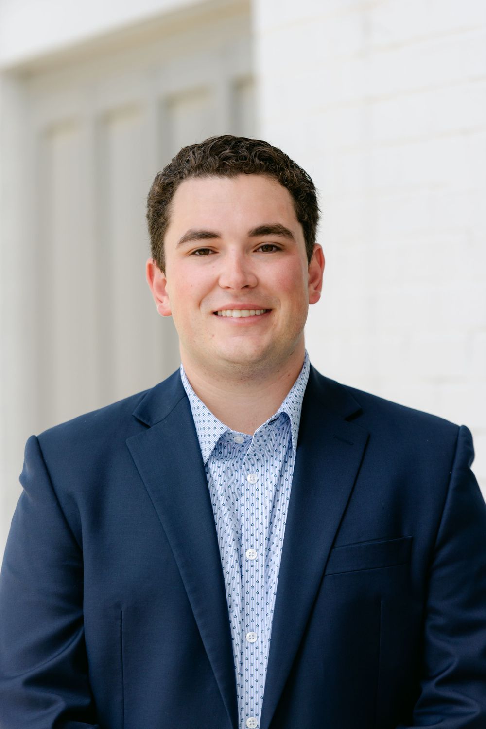 Professional headshot of a young man in a navy blazer smiling in front of a white building.