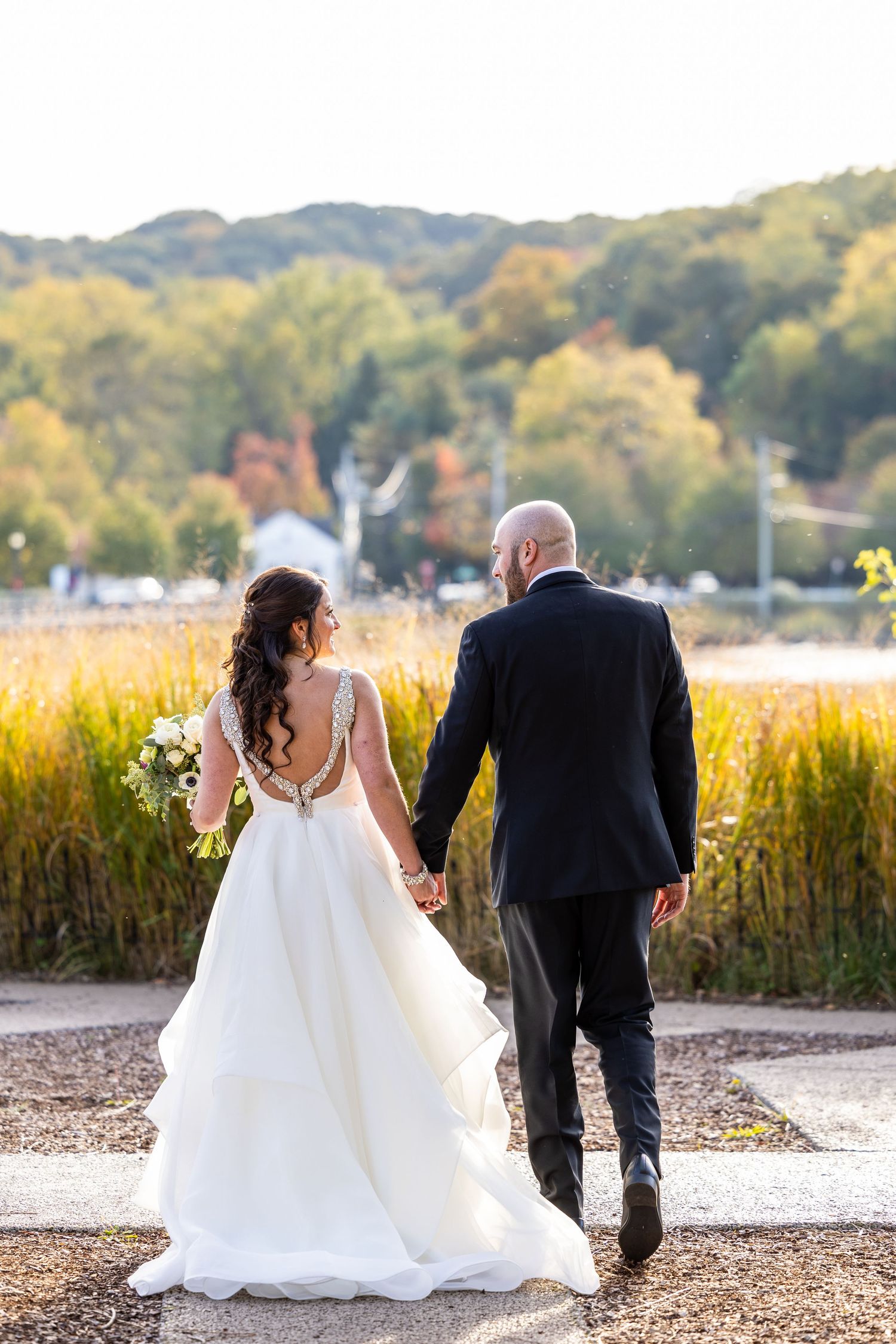 A bride and groom walking hand in hand through an autumn field at golden hour.