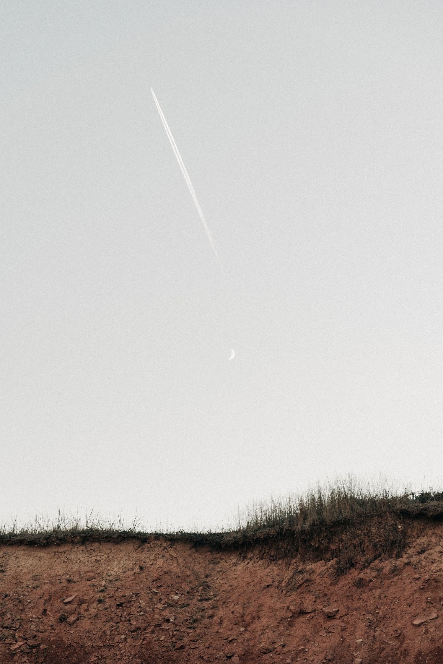 Minimalist landscape with contrail in grey sky over red earth and grass.