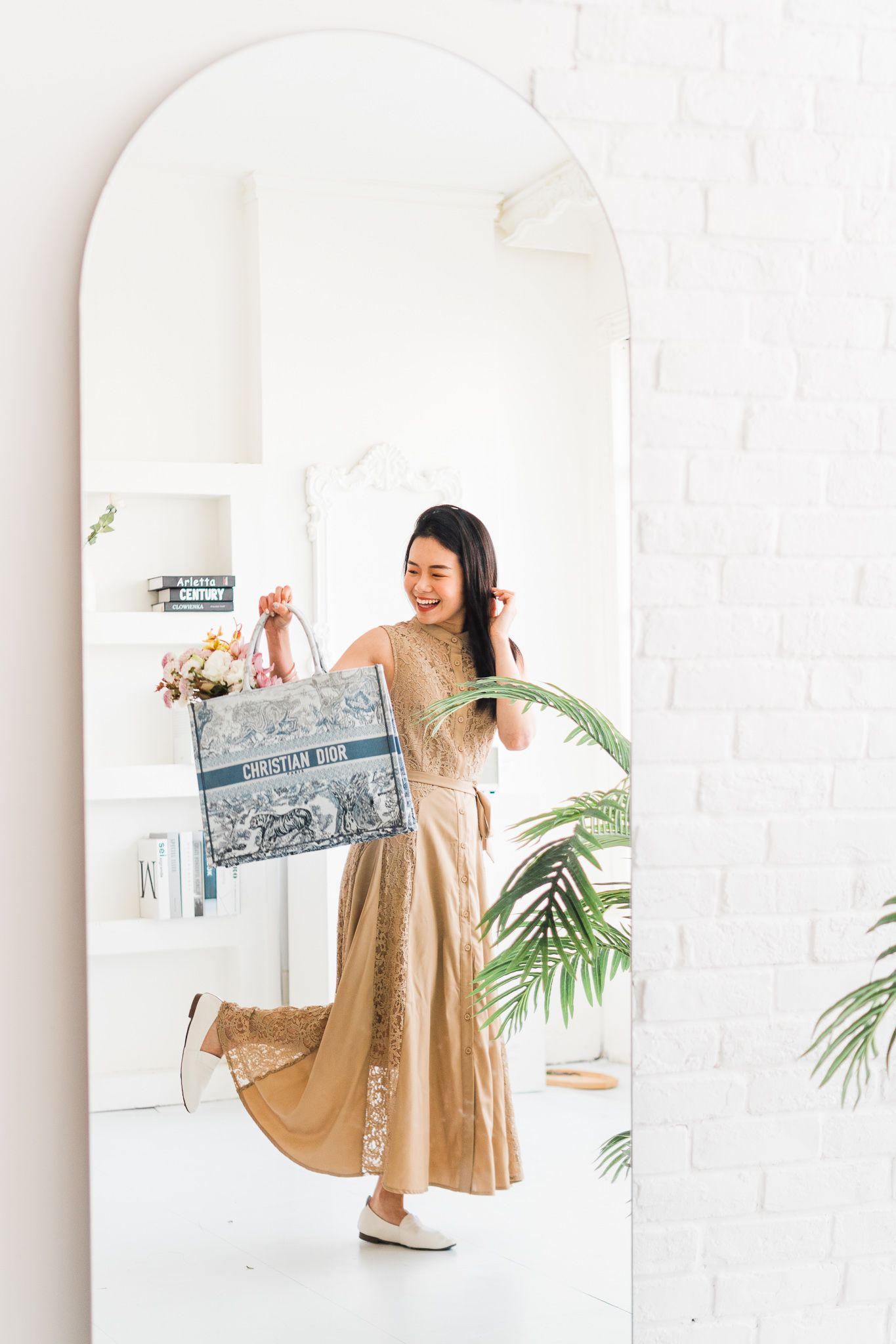 Woman in beige dress holding vintage artwork in front of arched mirror in bright white room.