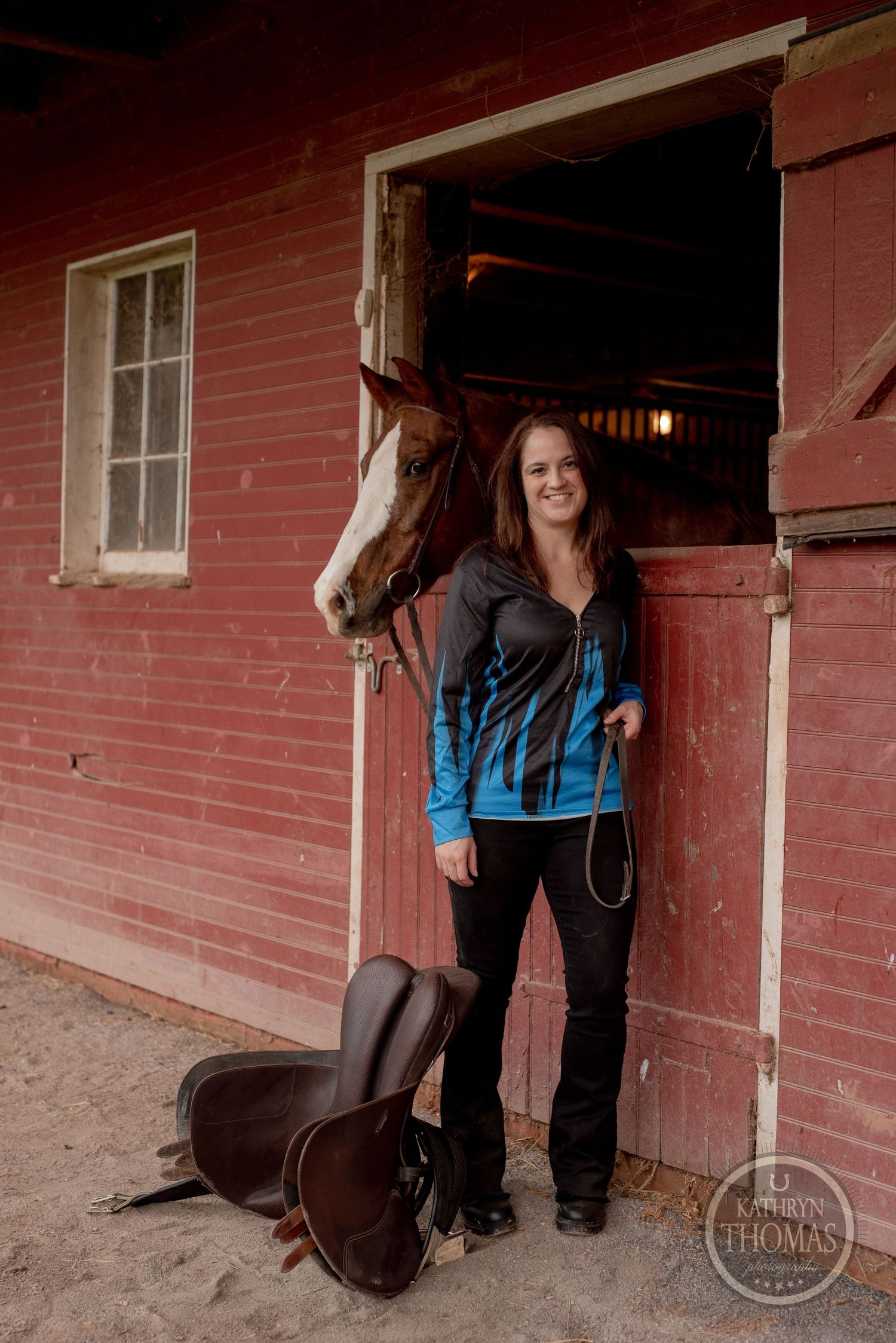 A chestnut horse peeks out of a red barn stable door with riding equipment visible.
