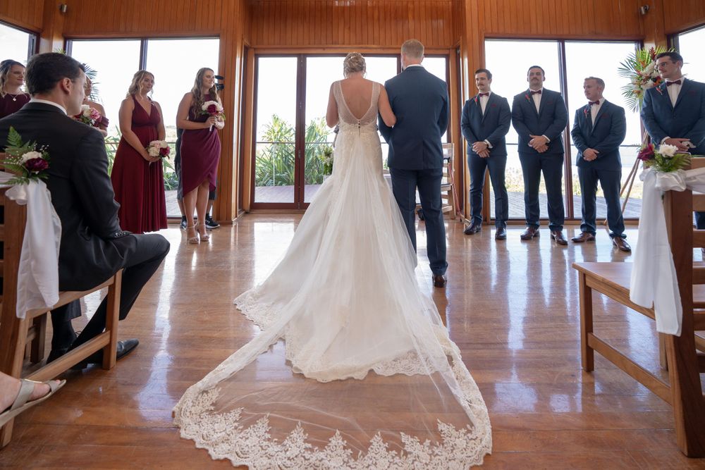 A couple stands at the altar during a wedding ceremony in a wooden interior venue with their wedding party in attendance.