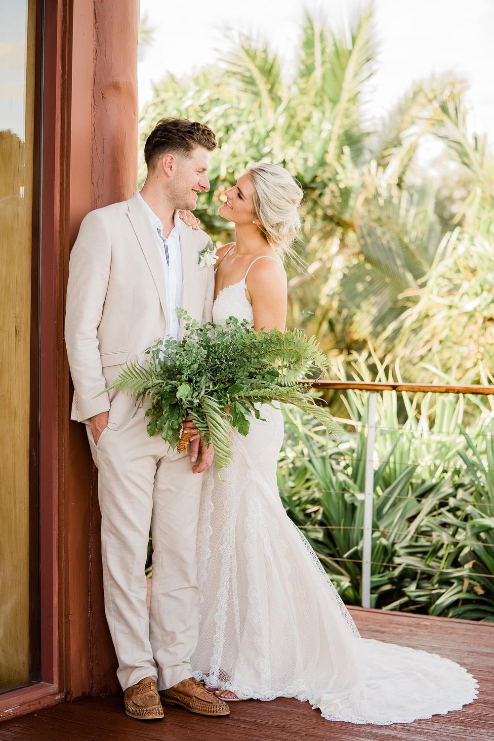 Couple in elegant white wedding attire share a joyful moment on a tropical balcony with palm trees in background.