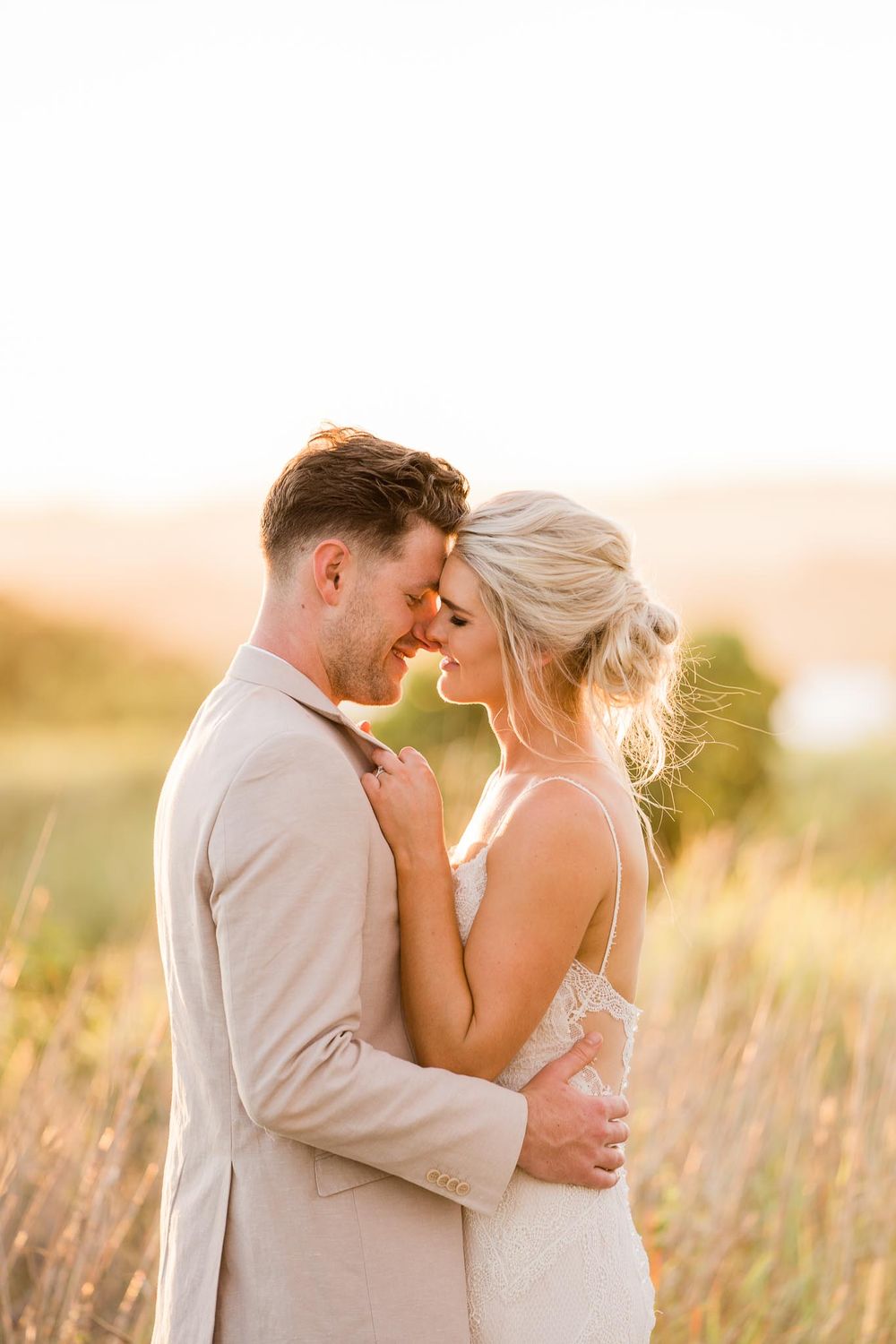 Romantic couple embraces in golden sunlit field during sunset wearing matching beige formal attire.