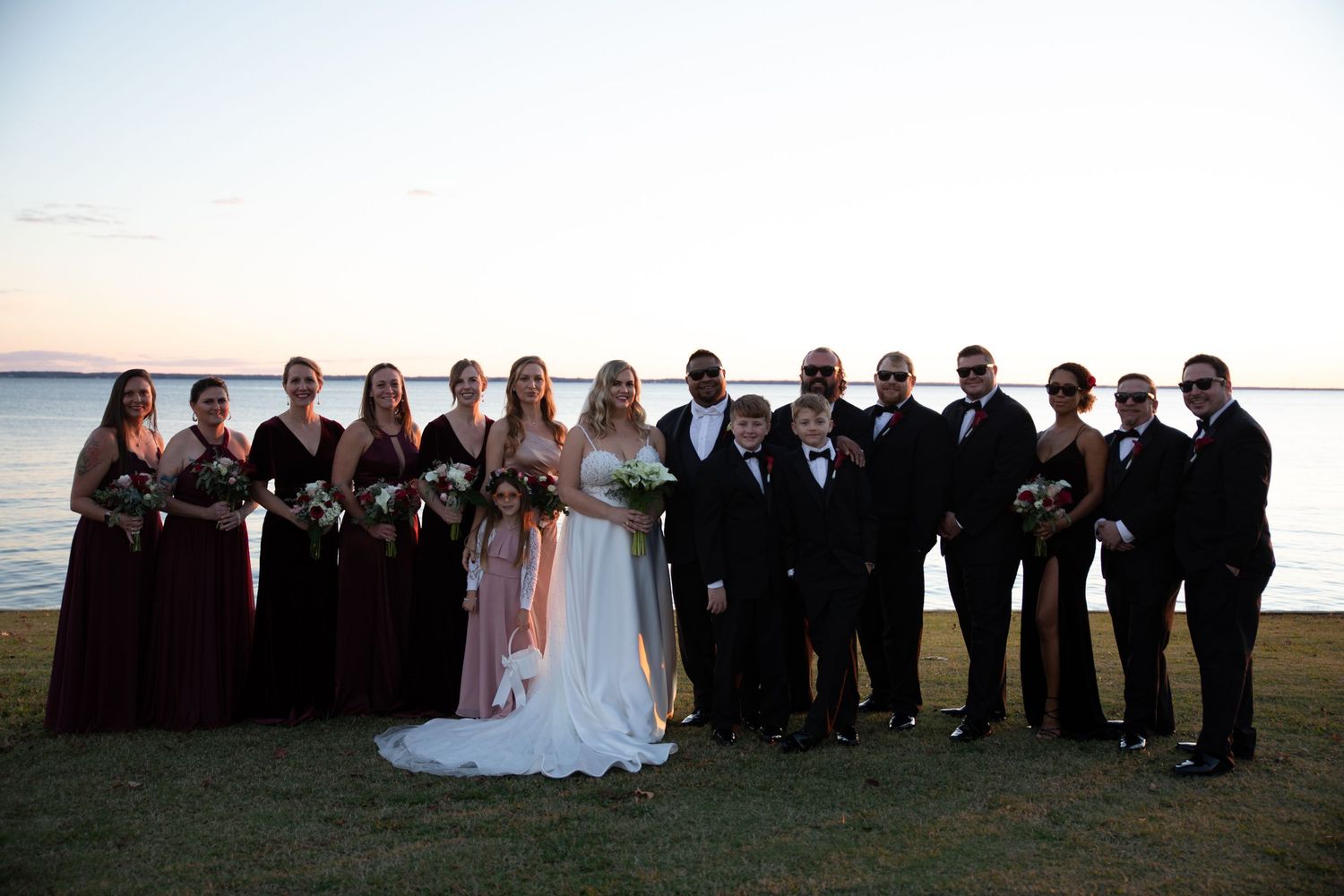 Wedding party lined up along waterfront at dusk wearing burgundy and black formal attire.