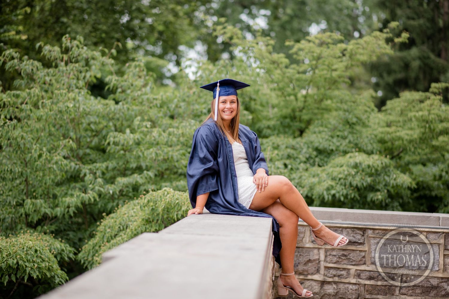 Graduate in blue regalia poses sitting on concrete wall with green foliage background.