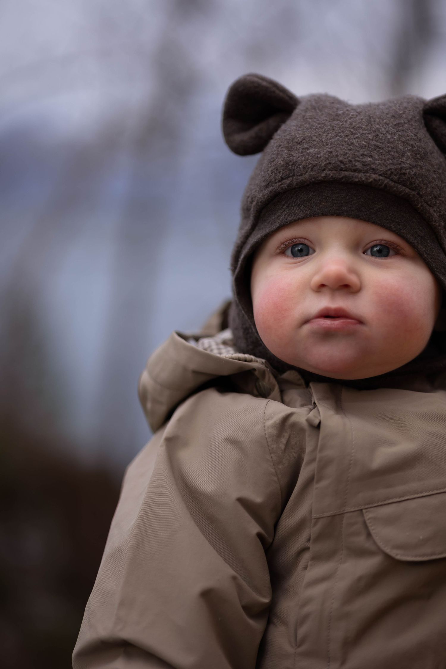 Small figure wearing brown bear hat and tan winter coat outdoors.