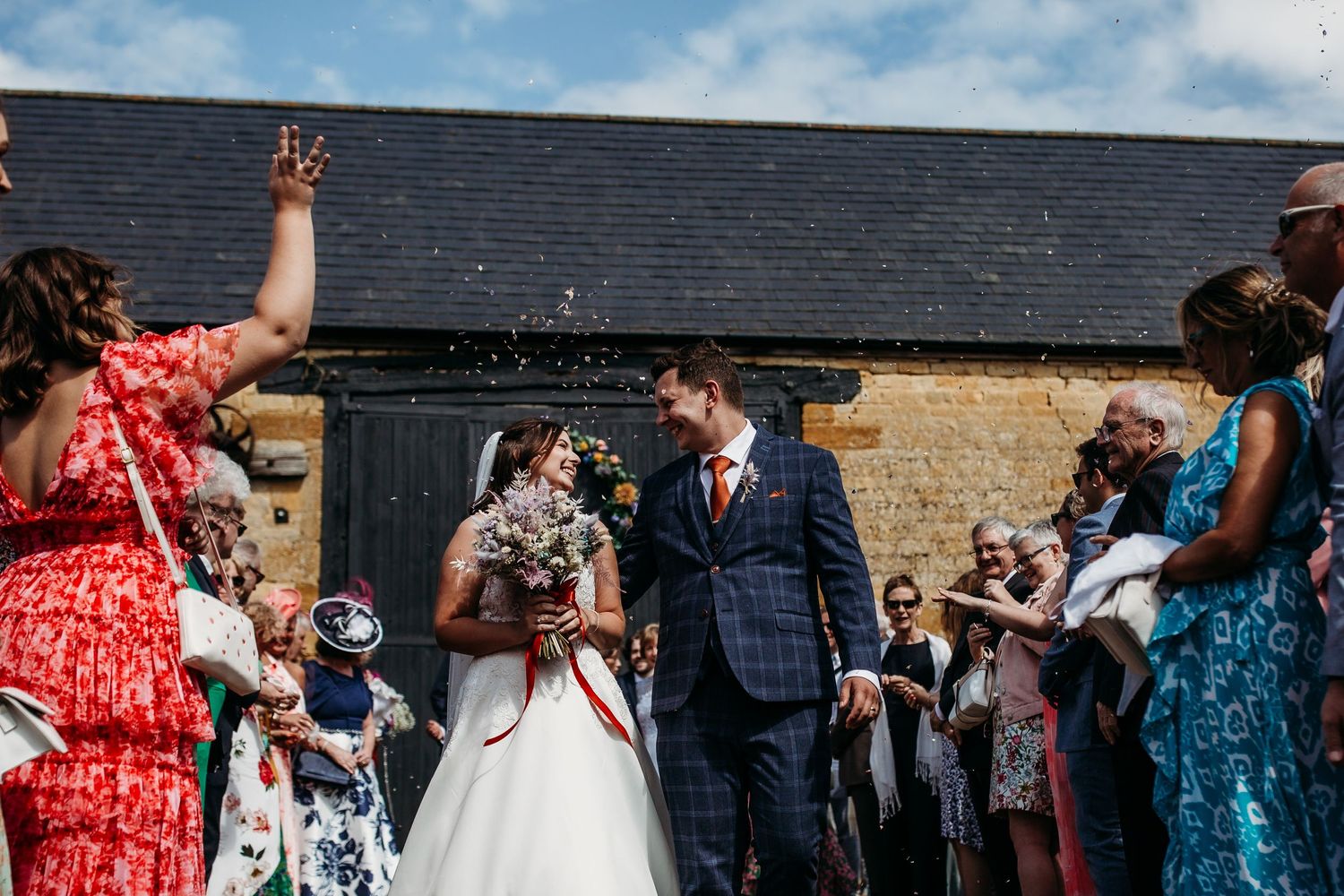Wedding party celebrates outside stone building under blue sky with guests throwing confetti.
