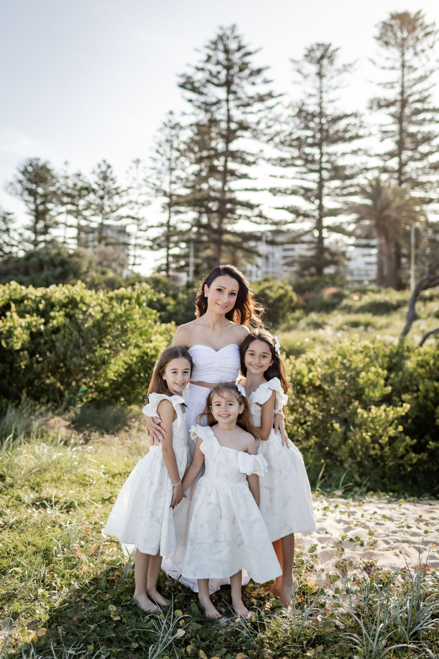 Group dressed in white posing together outdoors with tall pine trees and sunlight behind them.