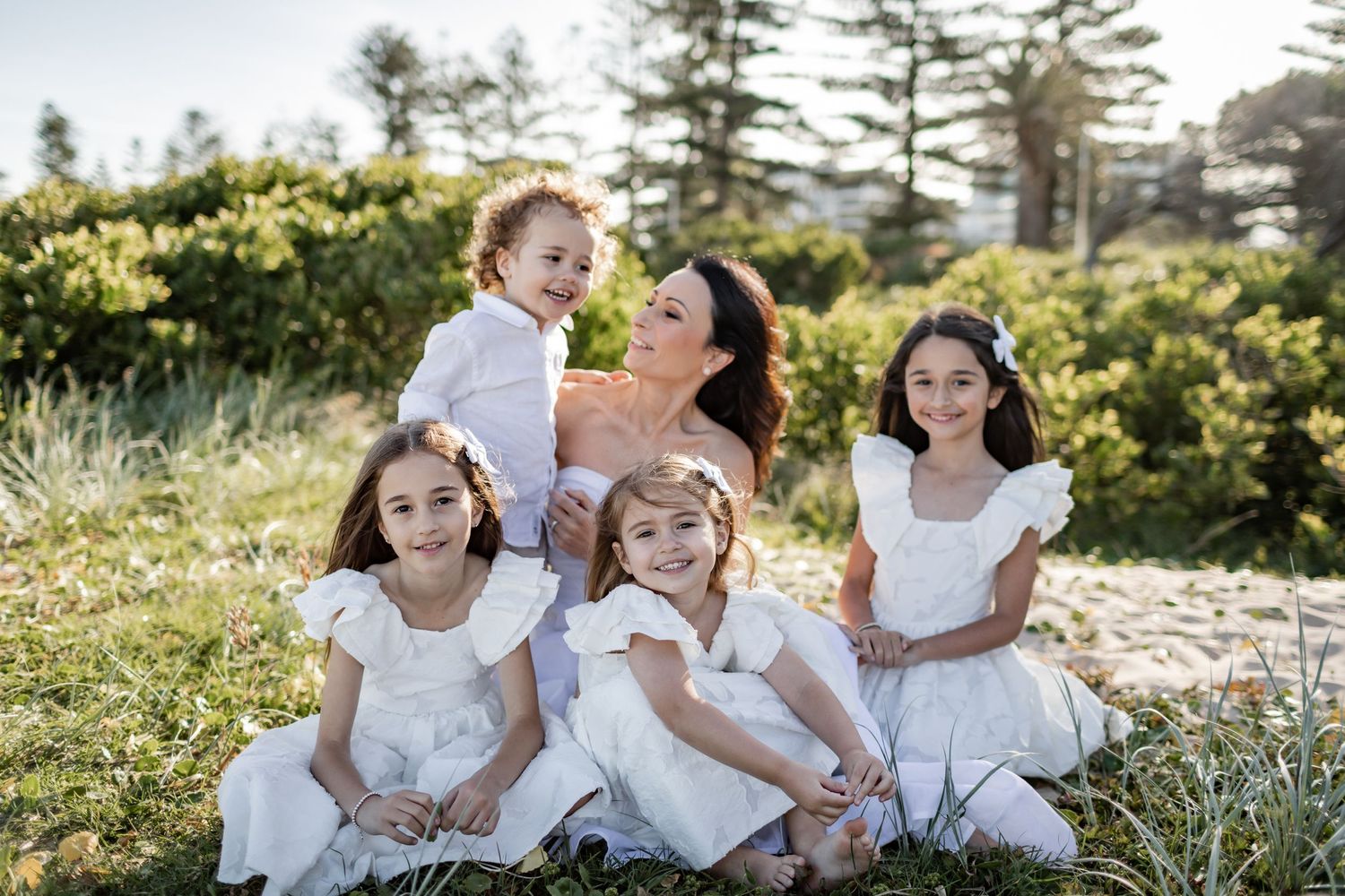 Group in white dresses sitting together on grassy hillside with pine trees in background.