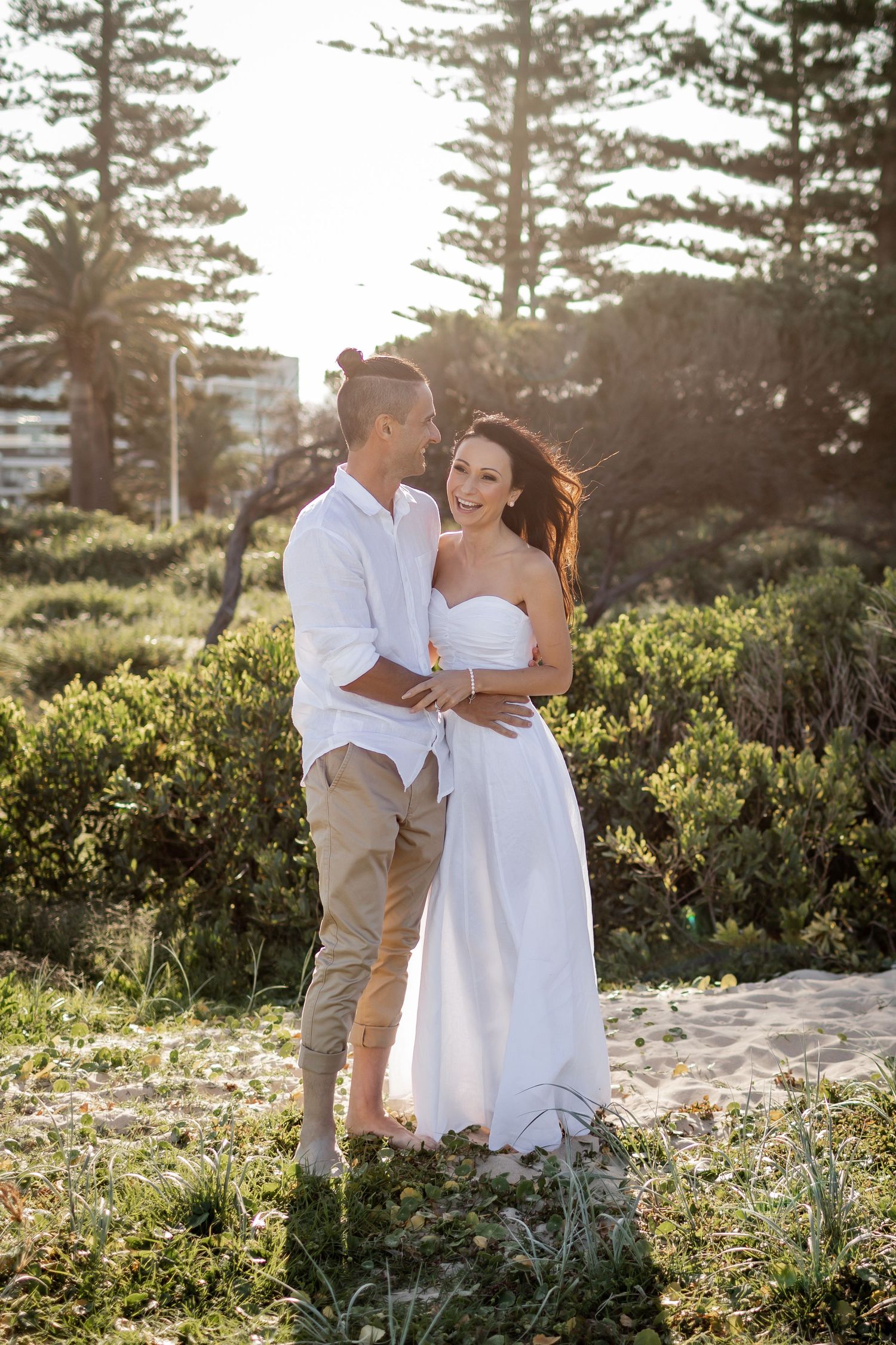 Couple in white embracing on coastal path during golden hour with pine trees in background.