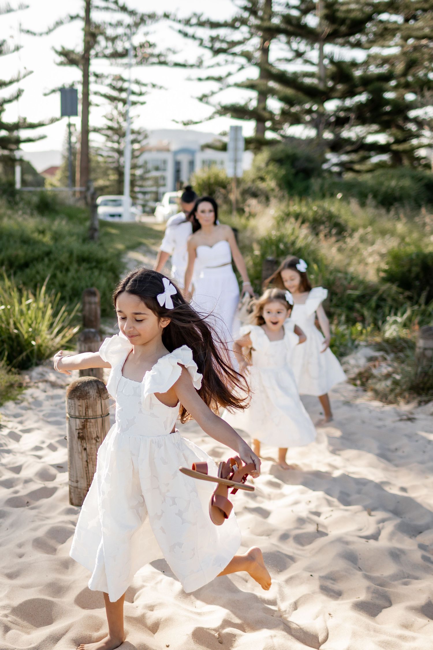 Children in white dresses run joyfully on a sandy beach path lined with pine trees.
