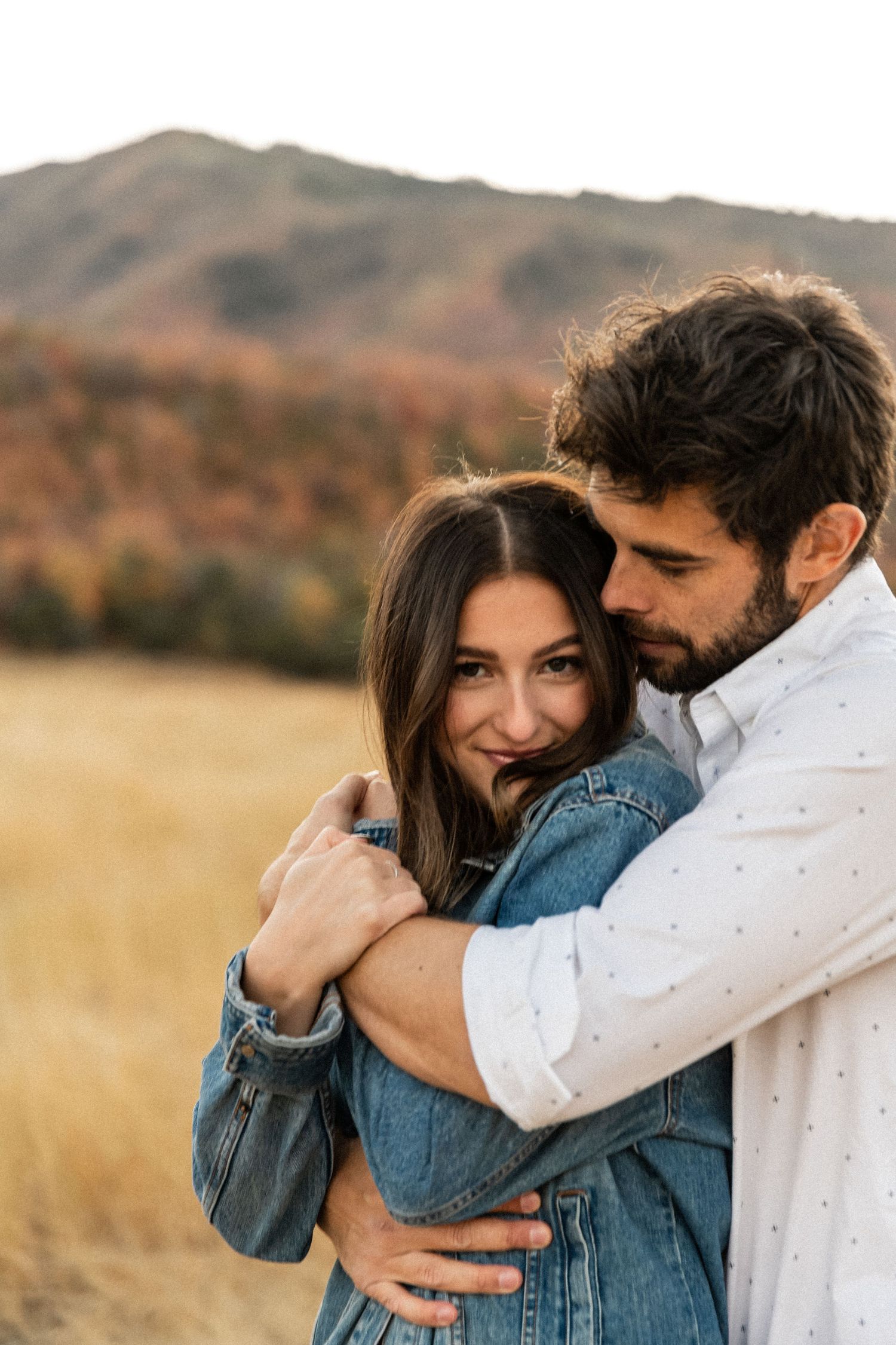 Couple embracing in a golden field with mountains in the background during sunset.