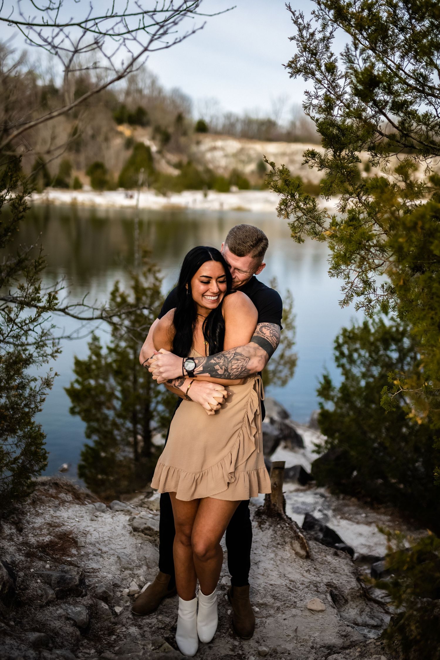 A playful couple enjoys a romantic photoshoot on rocky cliffs overlooking a peaceful lake.