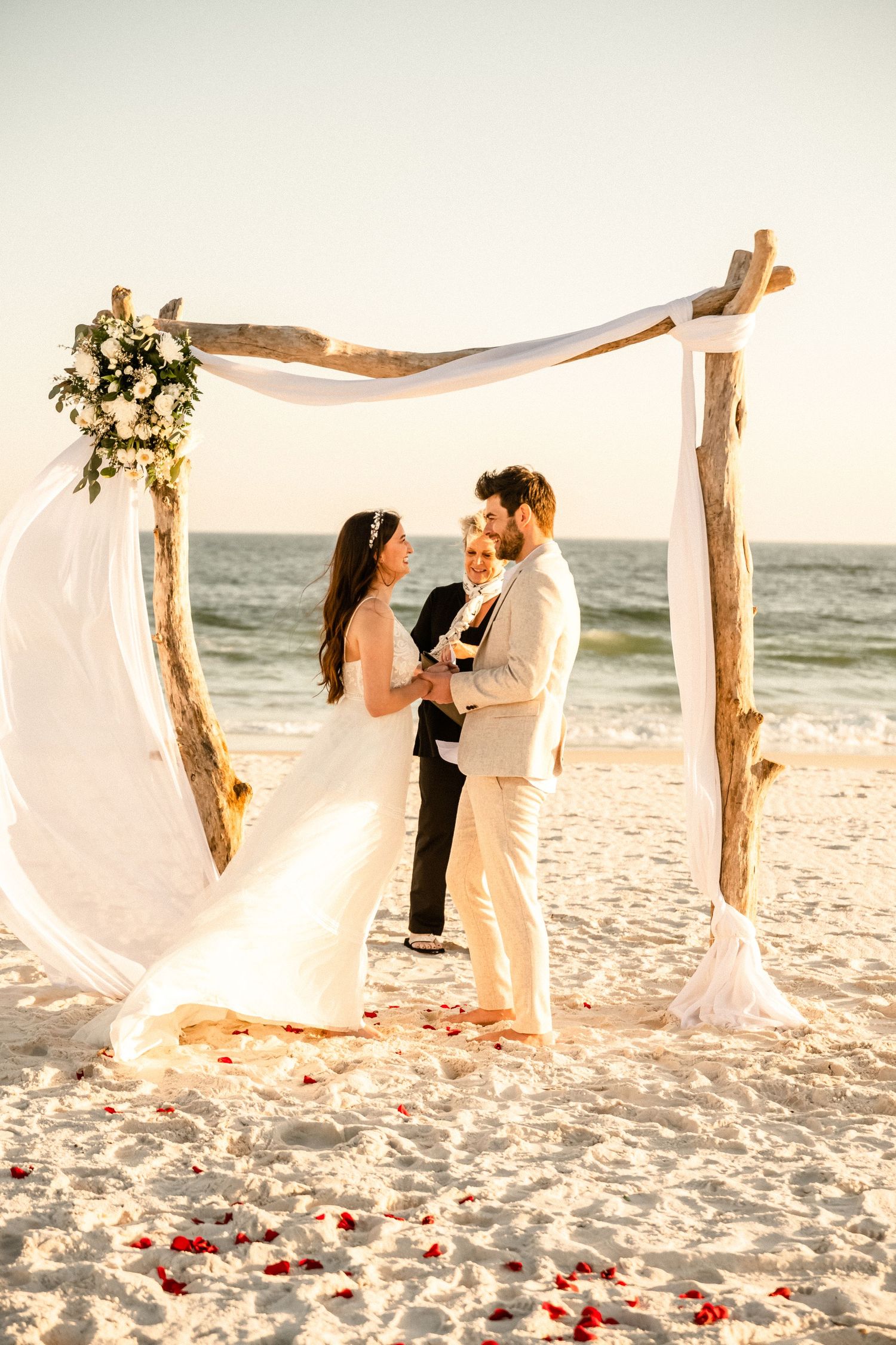 Beach wedding photos with wooden arch, flowing fabric, and rose petals on white sand.