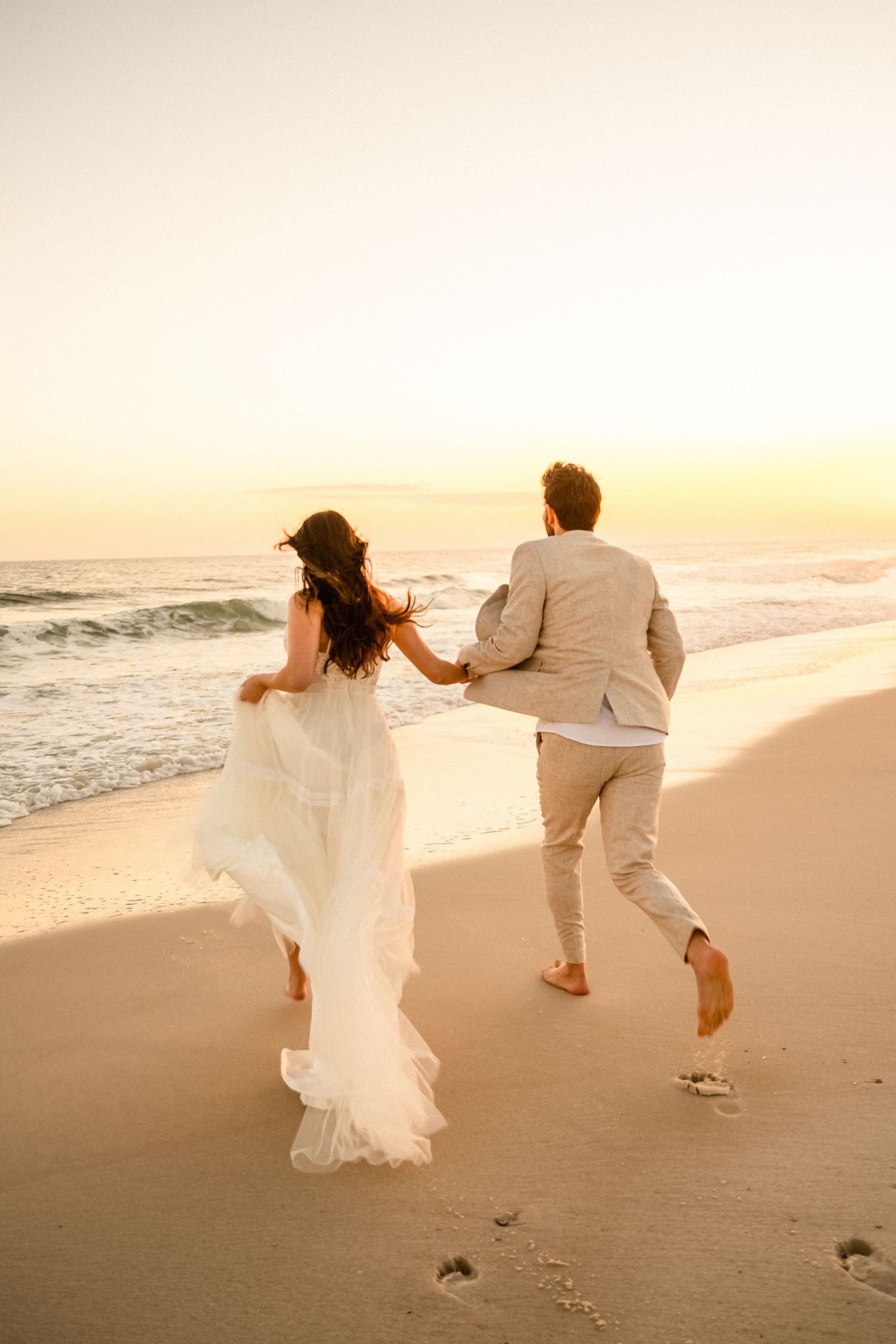 Couple running playfully on beach at sunset in white flowing outfits.