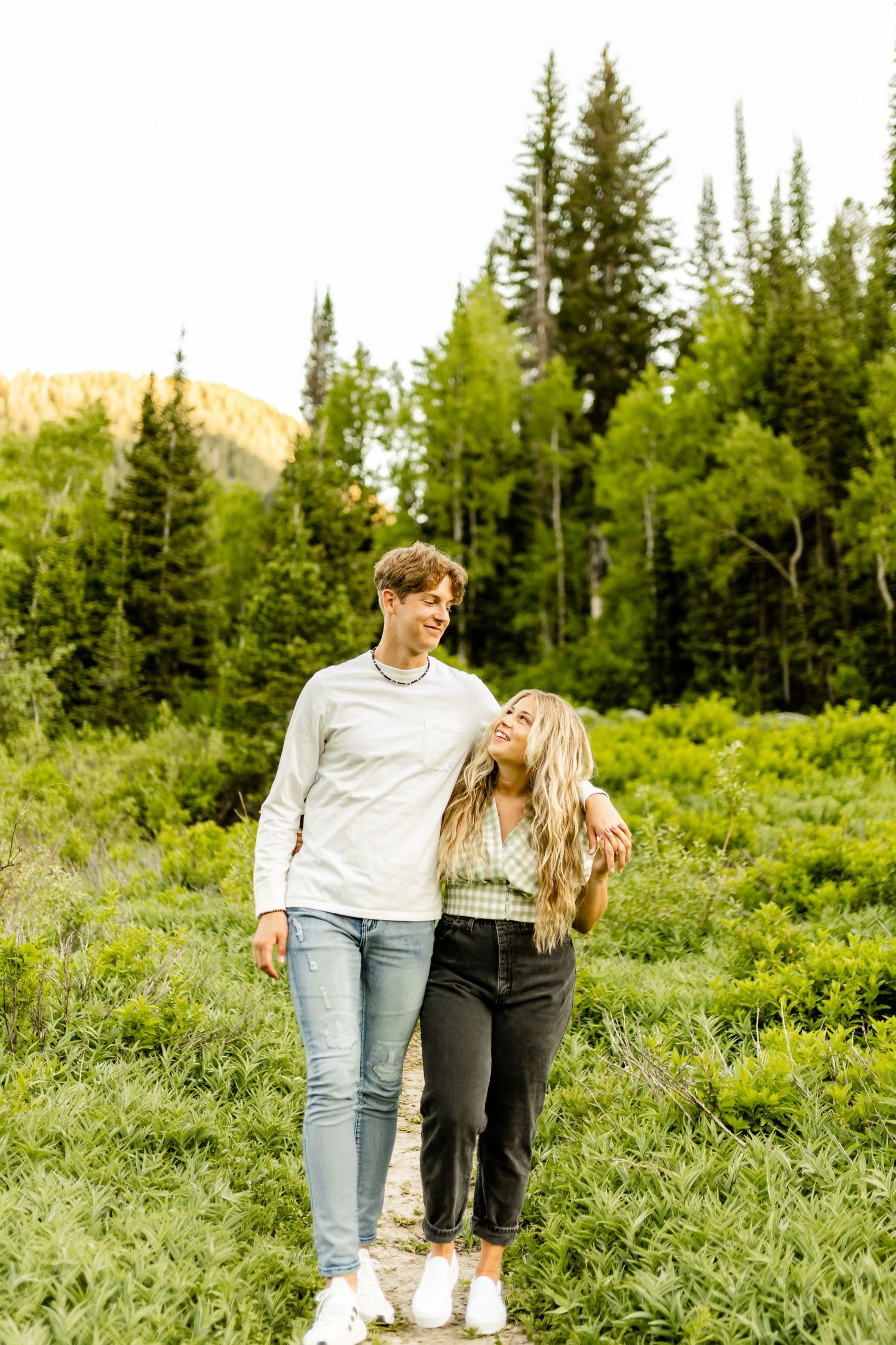 A couple shares romantic moments while walking through a scenic mountain meadow with evergreen trees.