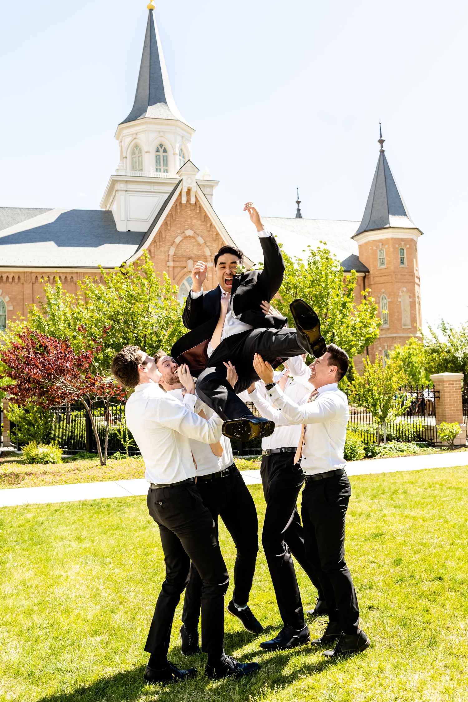 Groomsmen in black suits celebrate joyfully on a lawn with a church building in the background.