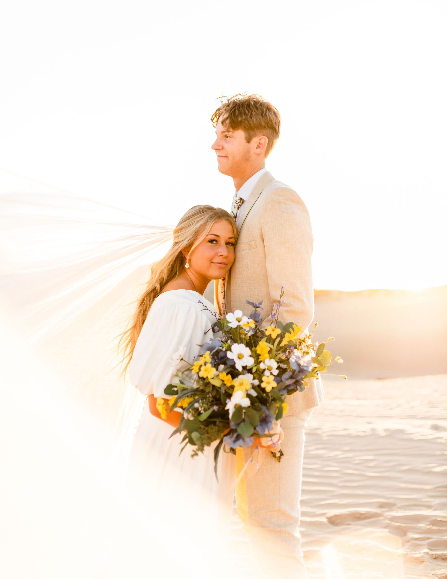 A couple embraces on a beach at sunset holding a bouquet of blue and yellow flowers.