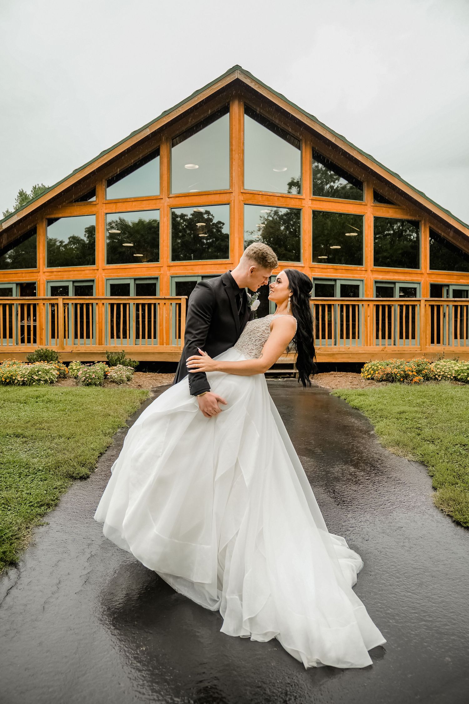 A romantic dip kiss in front of a rustic wooden lodge with large windows and timber beams.