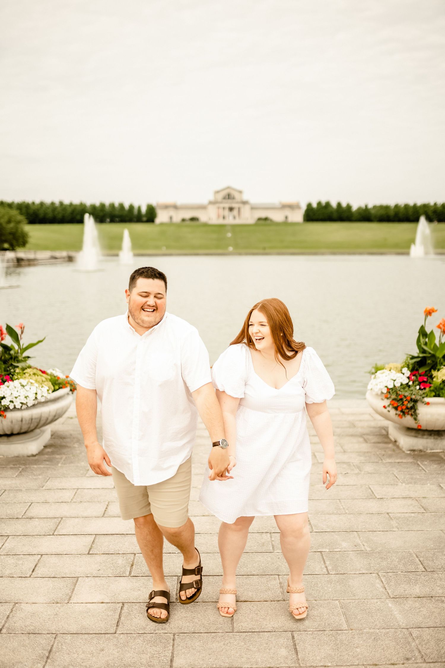 Playful couple in white outfits walking along a stone pathway near fountains.