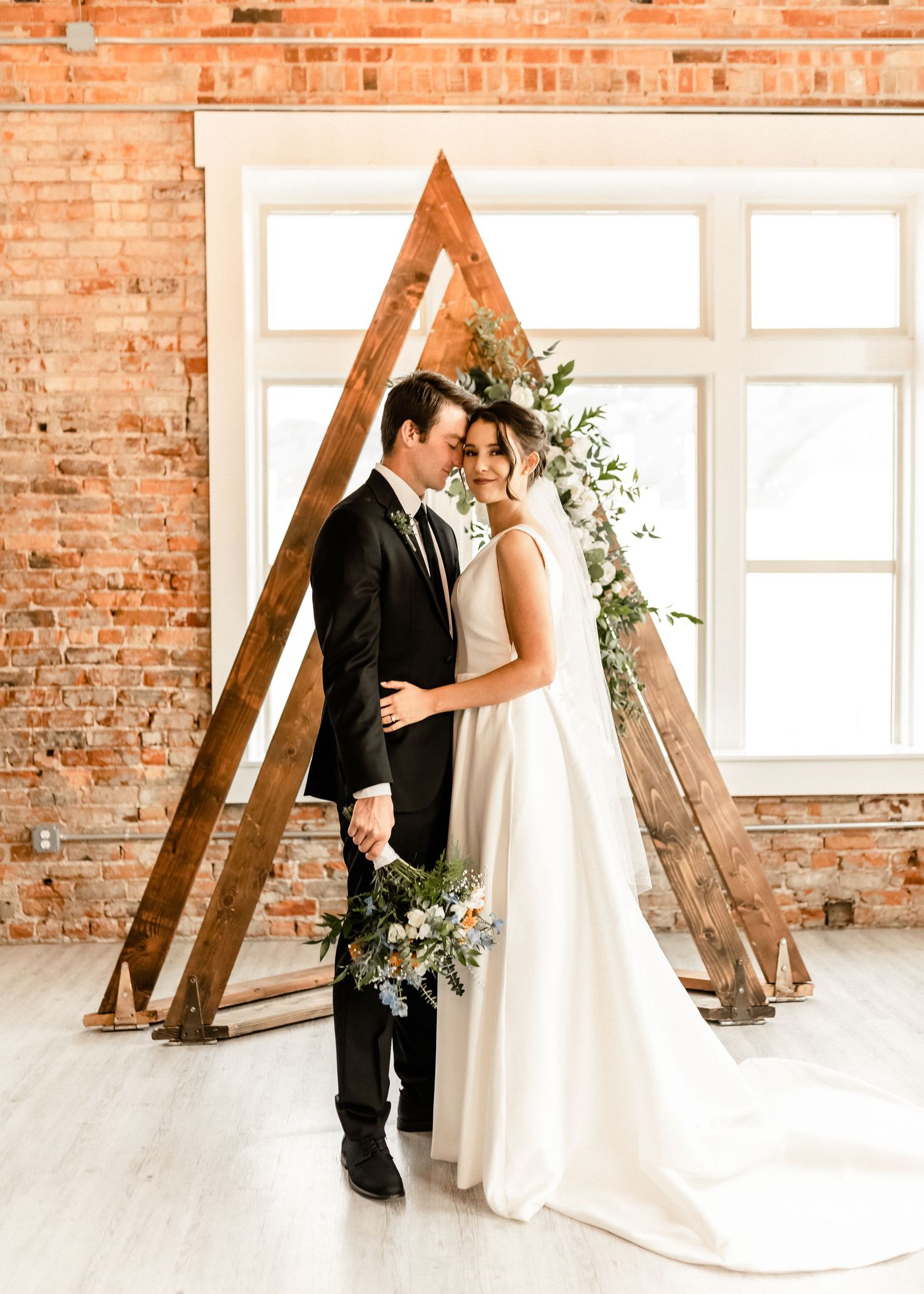 Wedding couple poses against rustic brick wall with wooden ladder and floral decorations.