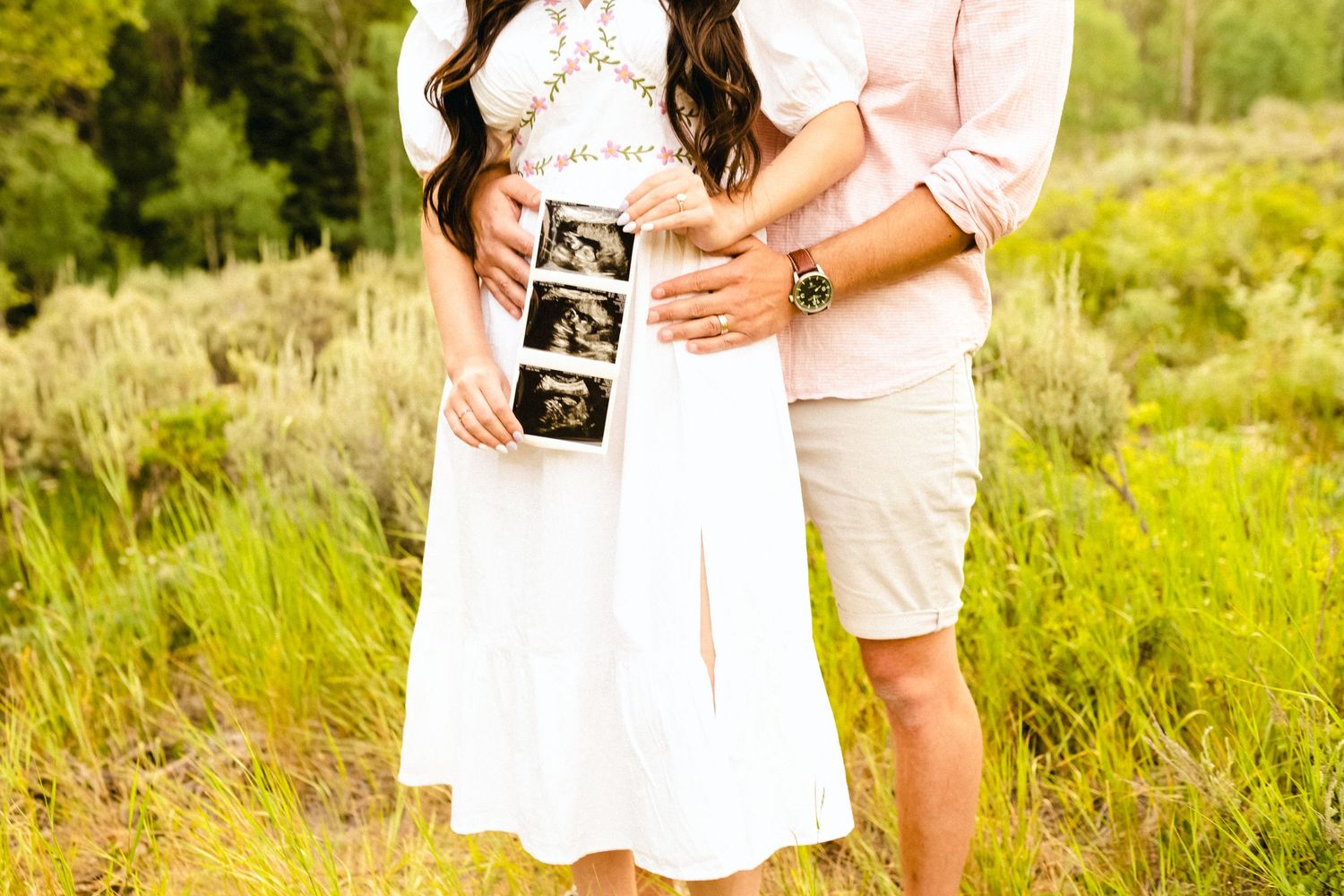 Couple holding ultrasound photos in a sunny outdoor field.