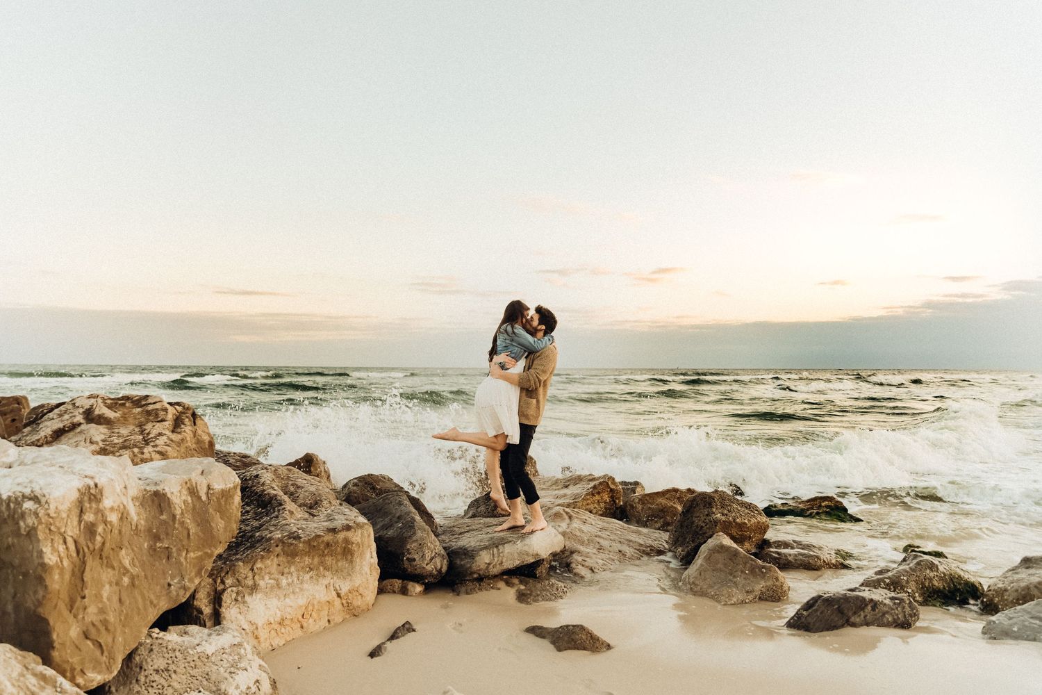 Coastal wedding photography session with waves crashing against rocky shore at sunset.