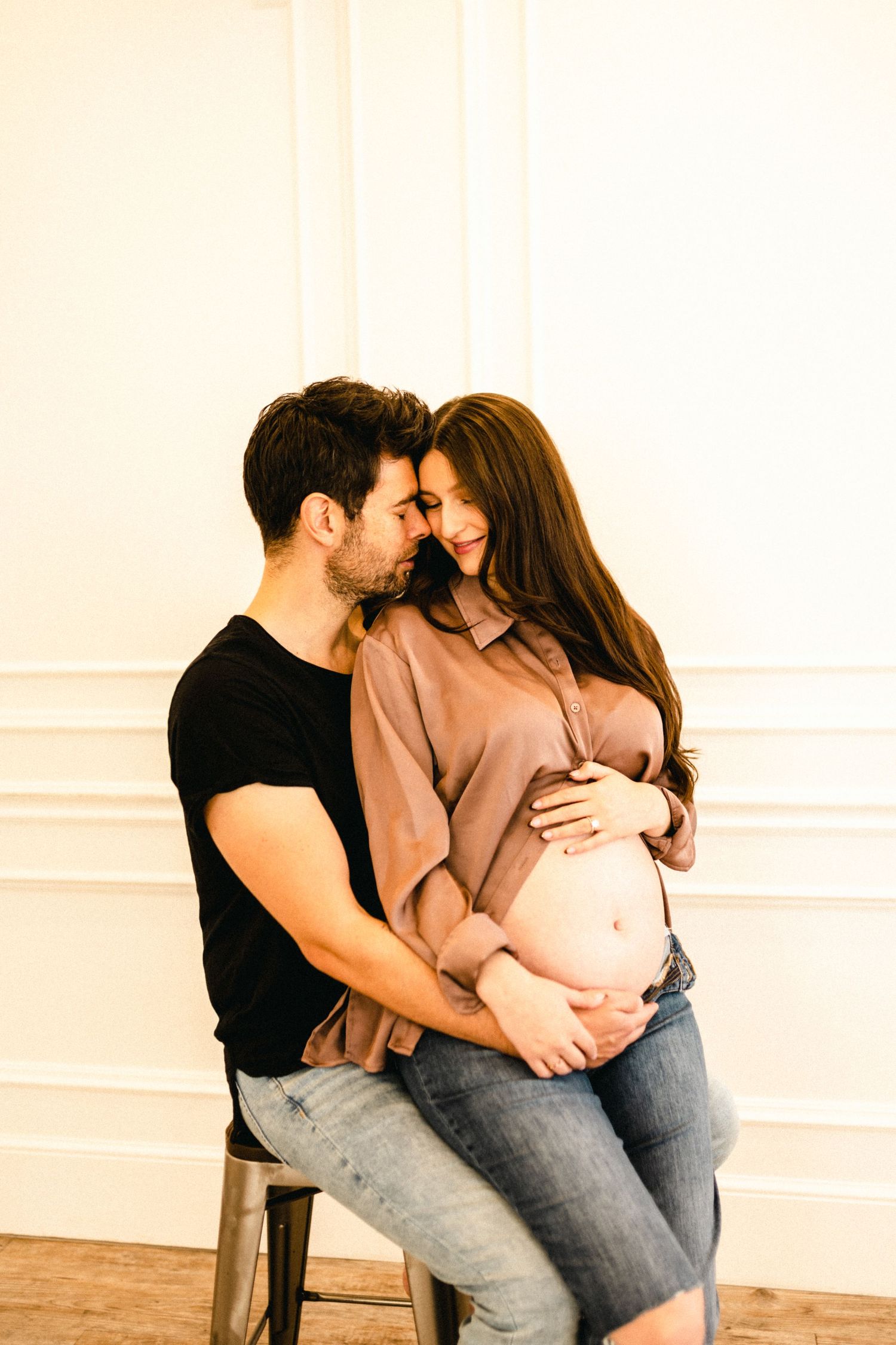 Intimate couple photoshoot showing tender moments in casual attire against a white wall.