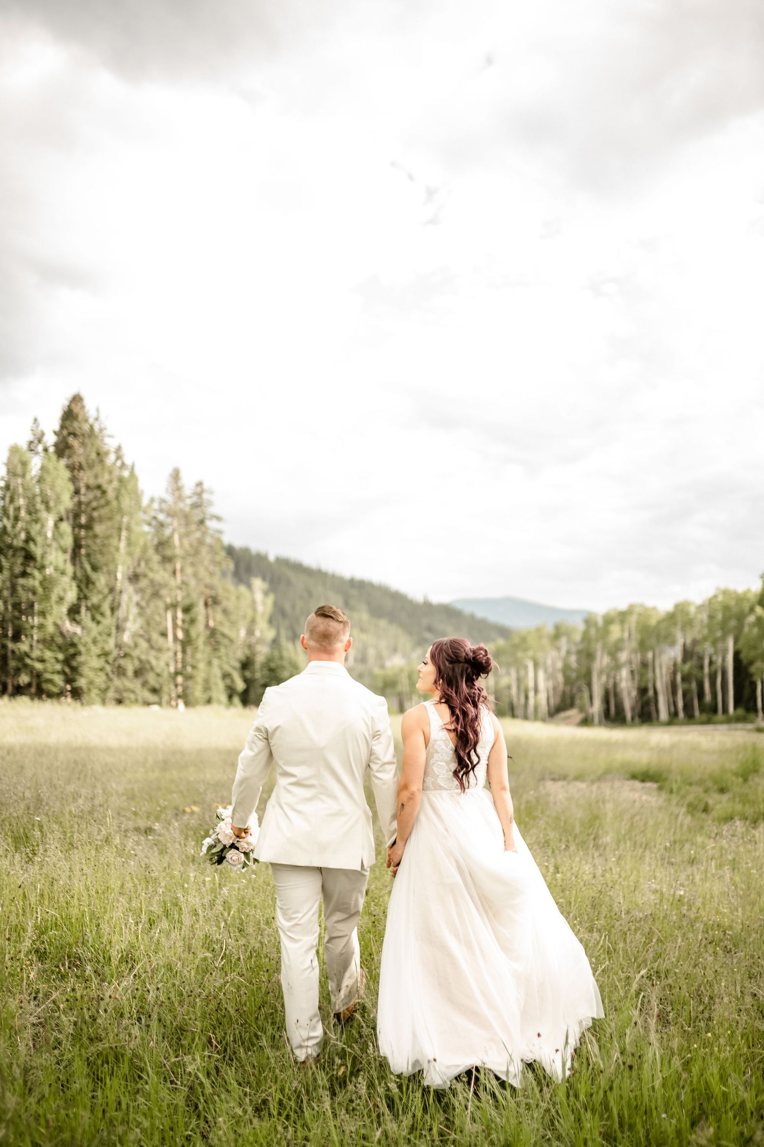 Couple walking through a scenic mountain meadow with tall grass and wildflowers.