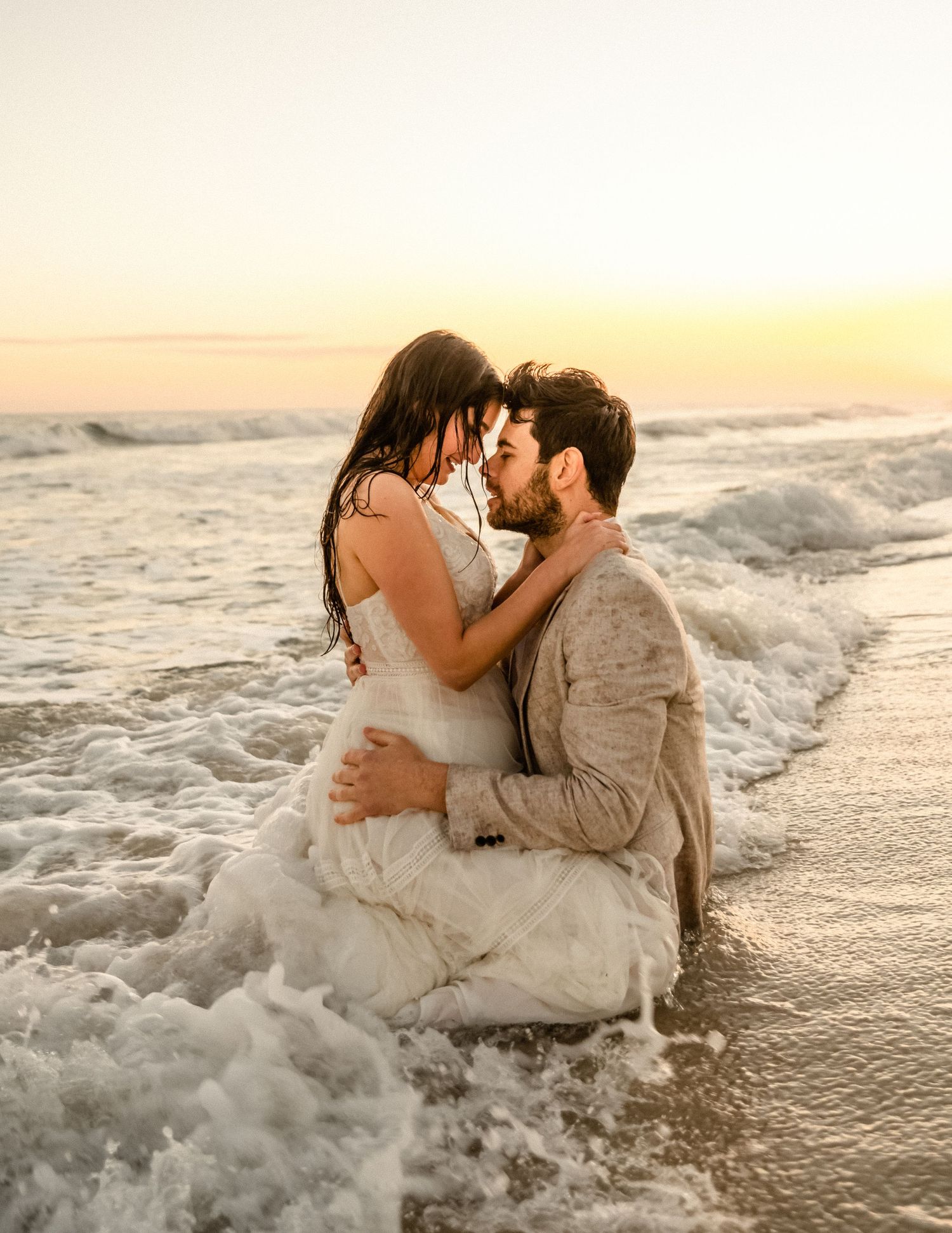 Romantic beach photoshoot with waves crashing around couple in formal attire.