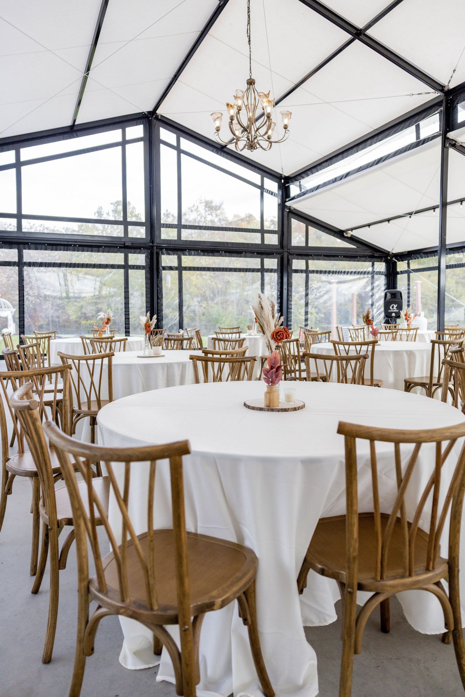 Elegant wedding reception venue with white round tables, wooden cross-back chairs, and a chandelier in a glass greenhouse.