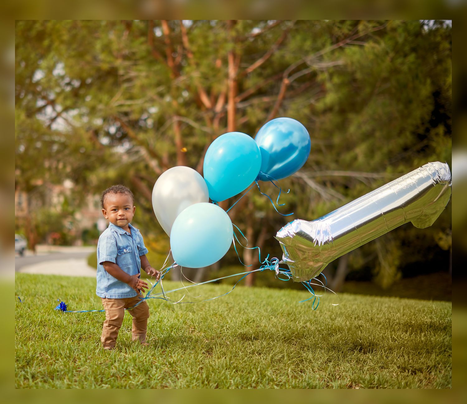 A small child holds blue and white balloons outdoors with trees in the background.