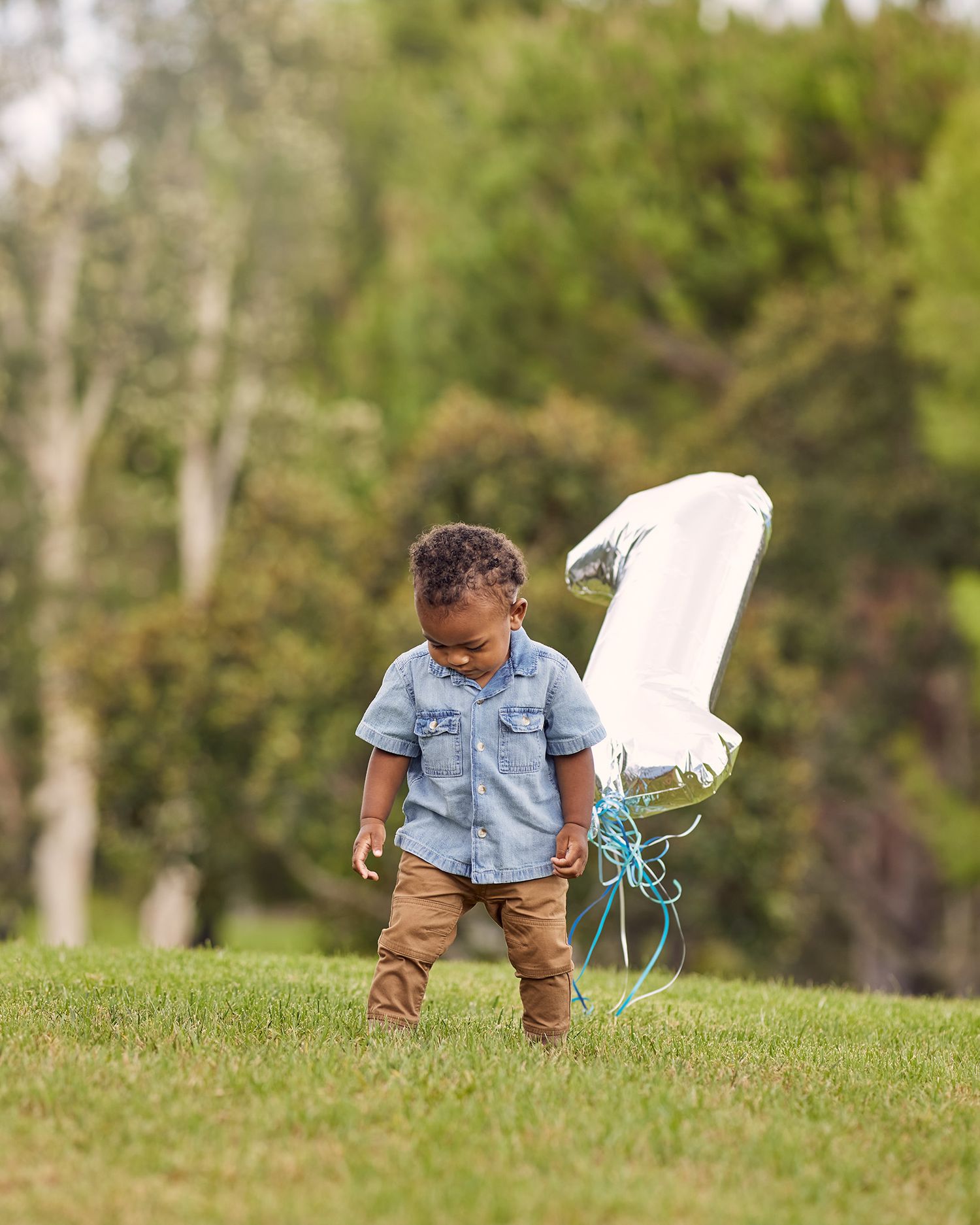 A toddler in a denim shirt stands on grass with a white balloon nearby.